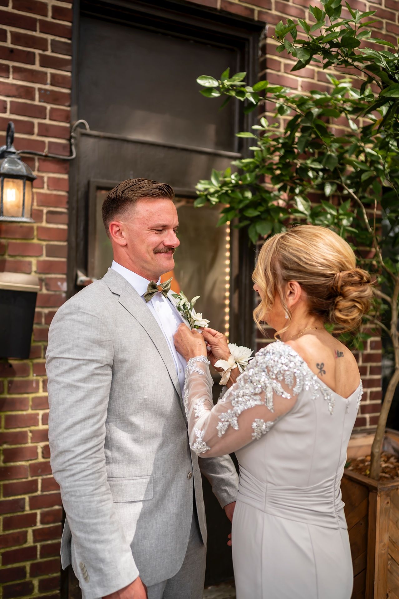 A touching wedding moment as someone pins a boutonniere on a groom wearing a light gray suit and bow tie.