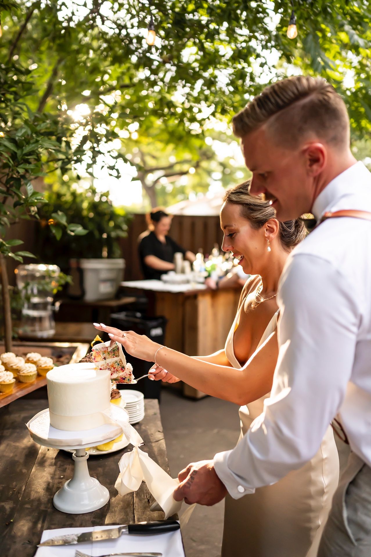 Couple checking phone while standing at rustic outdoor dessert table with white wedding cake and drinks at garden reception.
