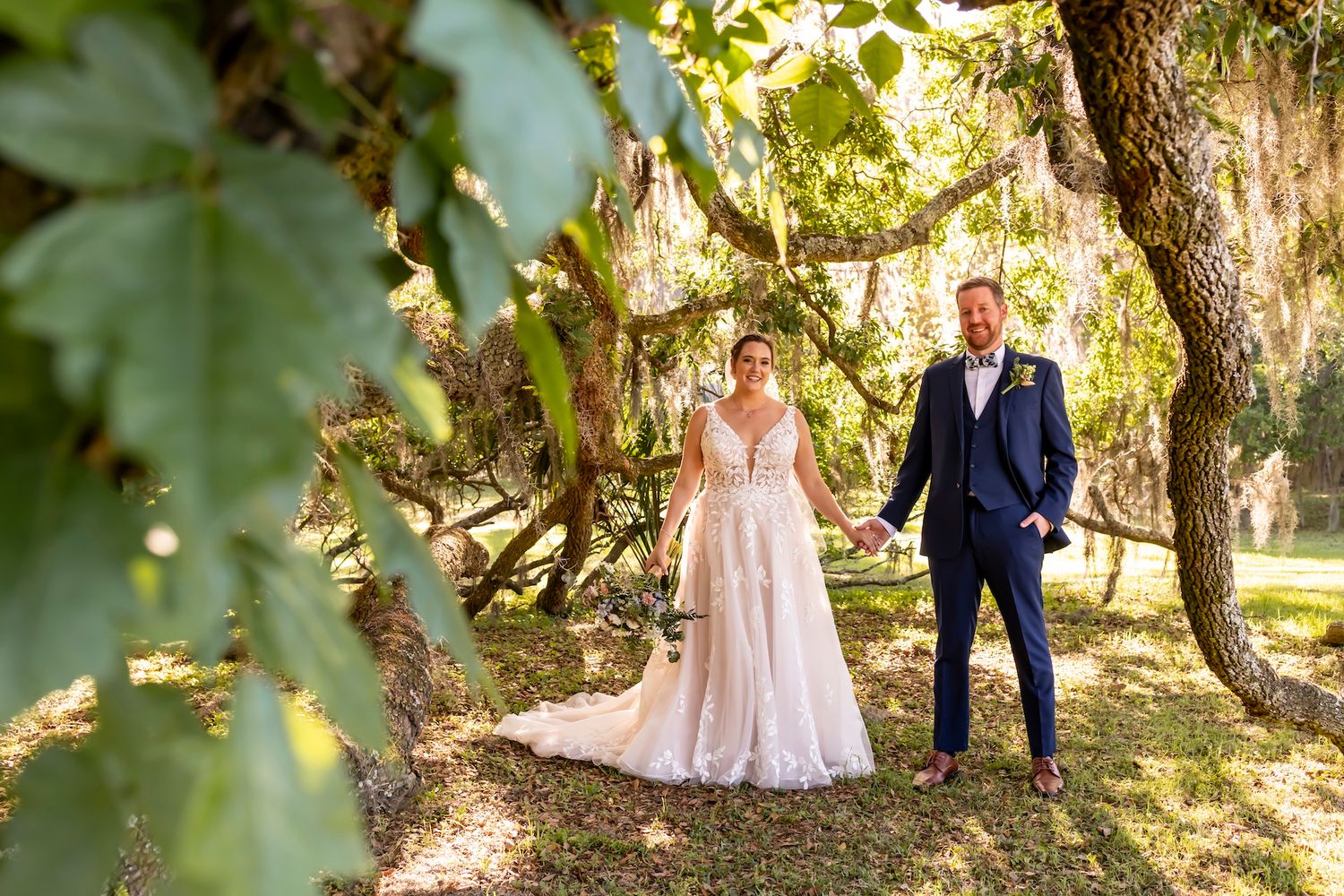 Elegant couple poses outdoors in garden setting with sunlit greenery and trees creating natural lighting.