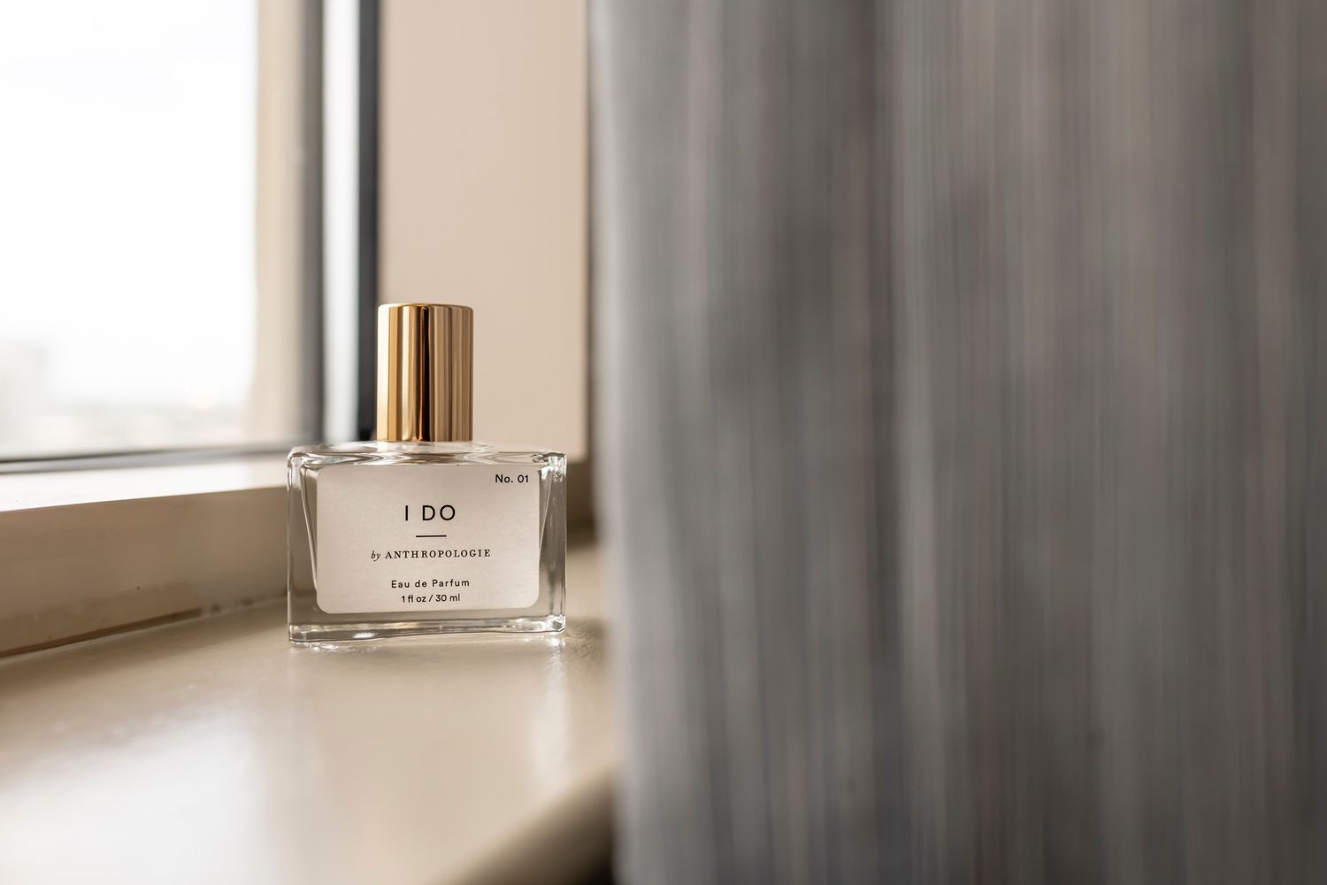 Clear glass perfume bottle with gold cap sits elegantly on white windowsill next to gray curtain in soft natural light.