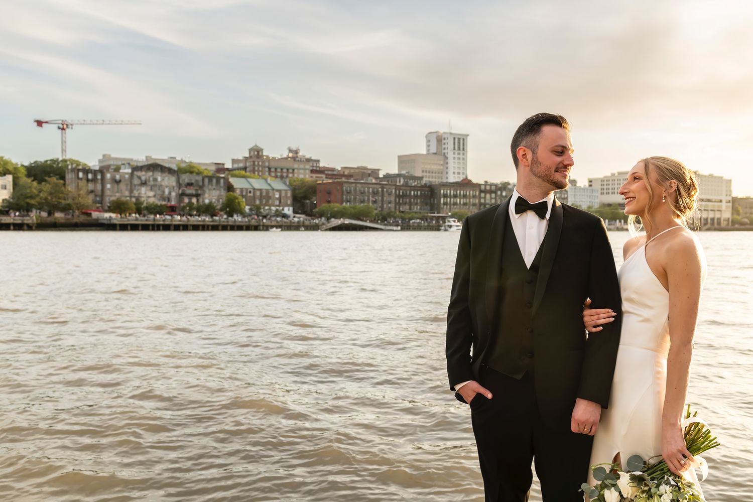 A romantic wedding couple stands together holding champagne glasses at a waterfront venue during sunset.