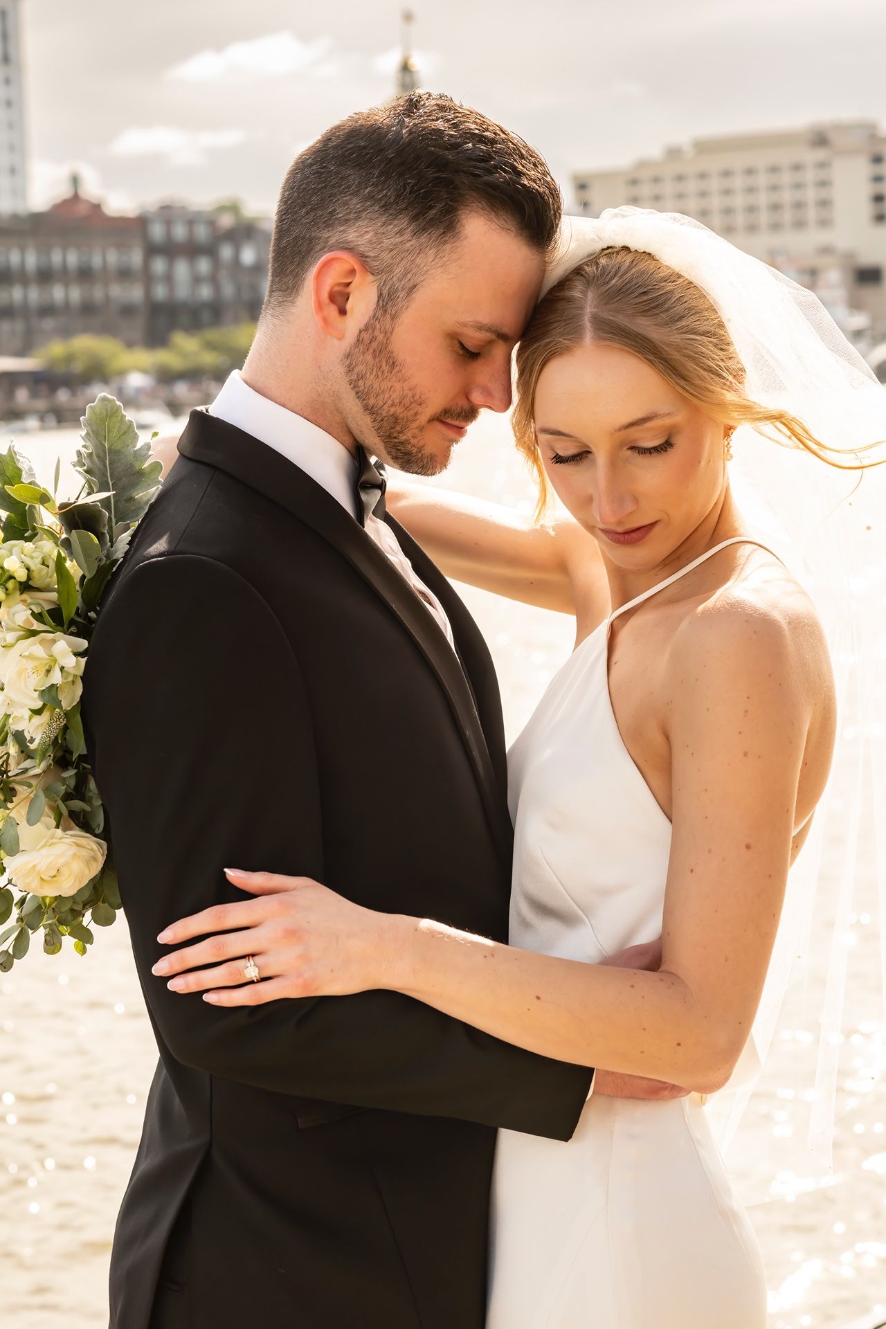 Newlywed couple in wedding attire embrace outdoors in warm sunlight with cityscape backdrop and white floral bouquet.