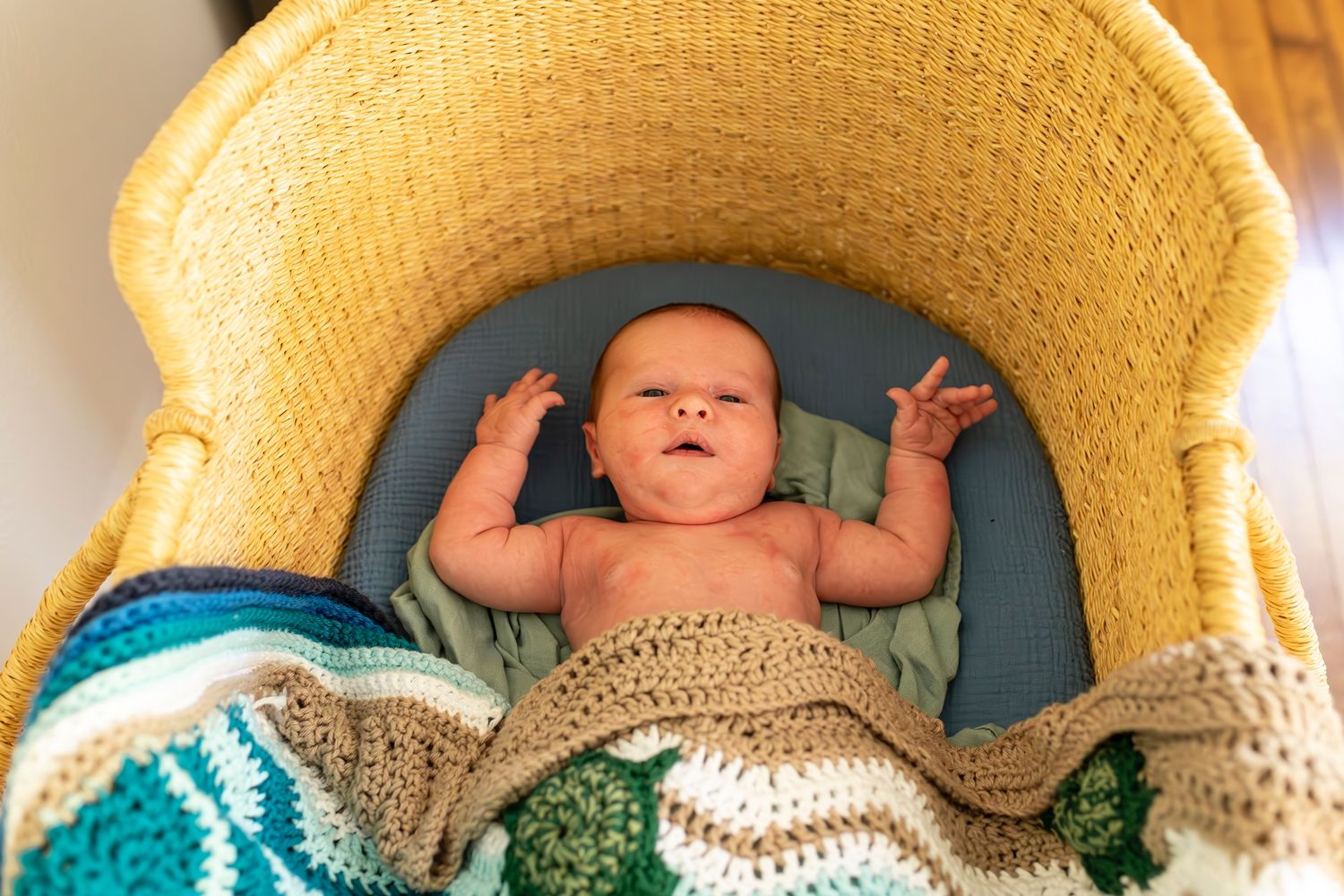 A baby lies in a yellow bassinet covered with a handmade blue and beige crocheted blanket while reaching up.