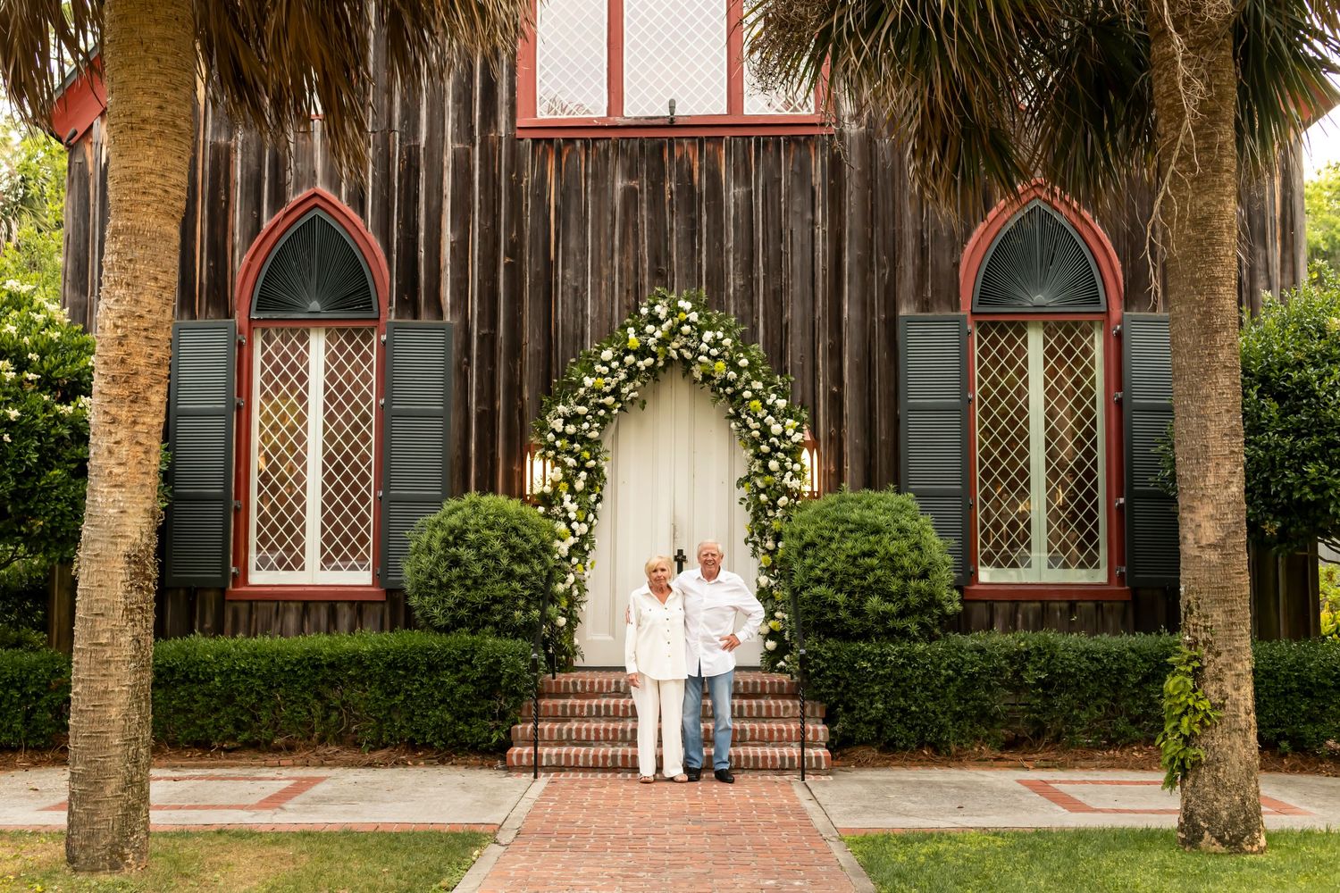 A rustic wooden church exterior with arched windows, palm trees, and a floral archway on the entrance steps.