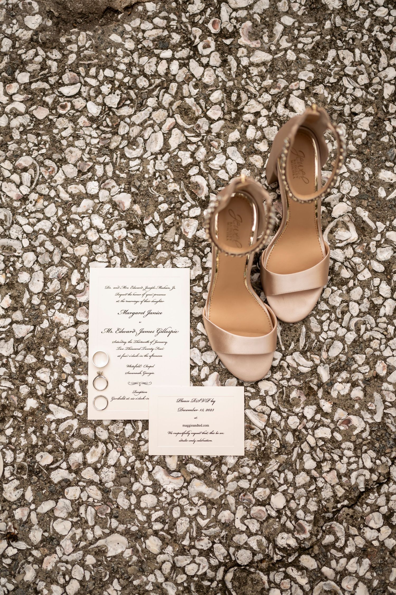 Elegant beige wedding shoes and invitation cards arranged on a textured stone surface with scattered ring details.