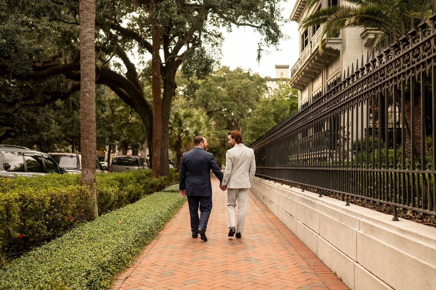 Two grooms stroll along a tree-lined path with wrought iron fencing in Historic Savannah.