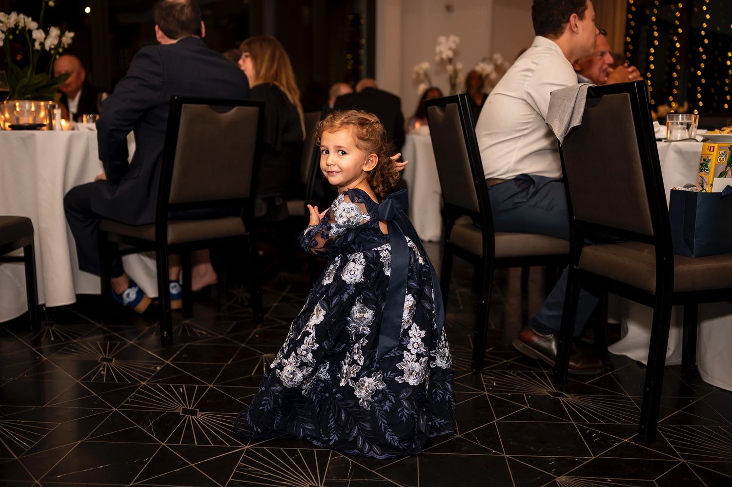 Young child in floral navy dress twirls on dance floor at elegant reception with tables and string lights in background.