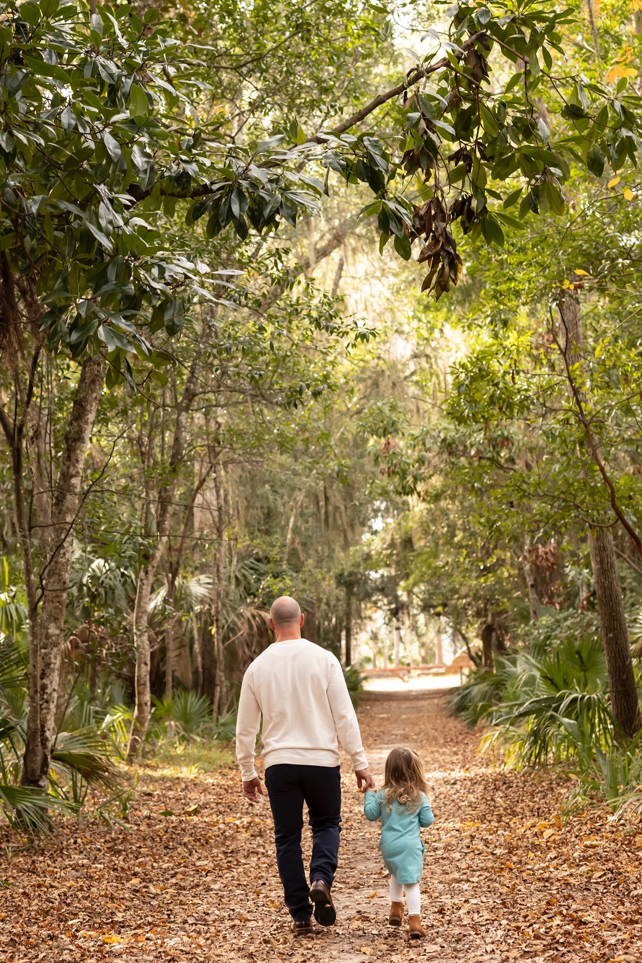 A parent and small child walk together on a tree-lined nature trail covered in fallen autumn leaves.