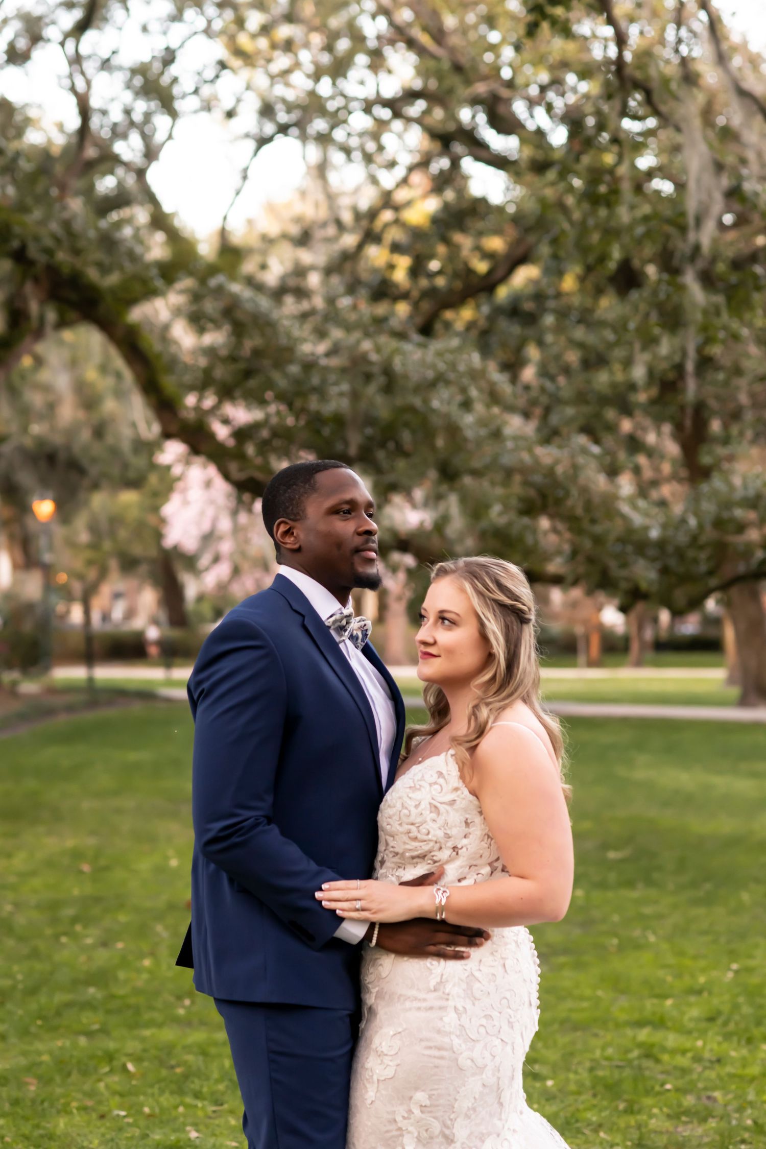 A couple in formal wedding attire embrace outdoors under oak trees at sunset in a garden setting.