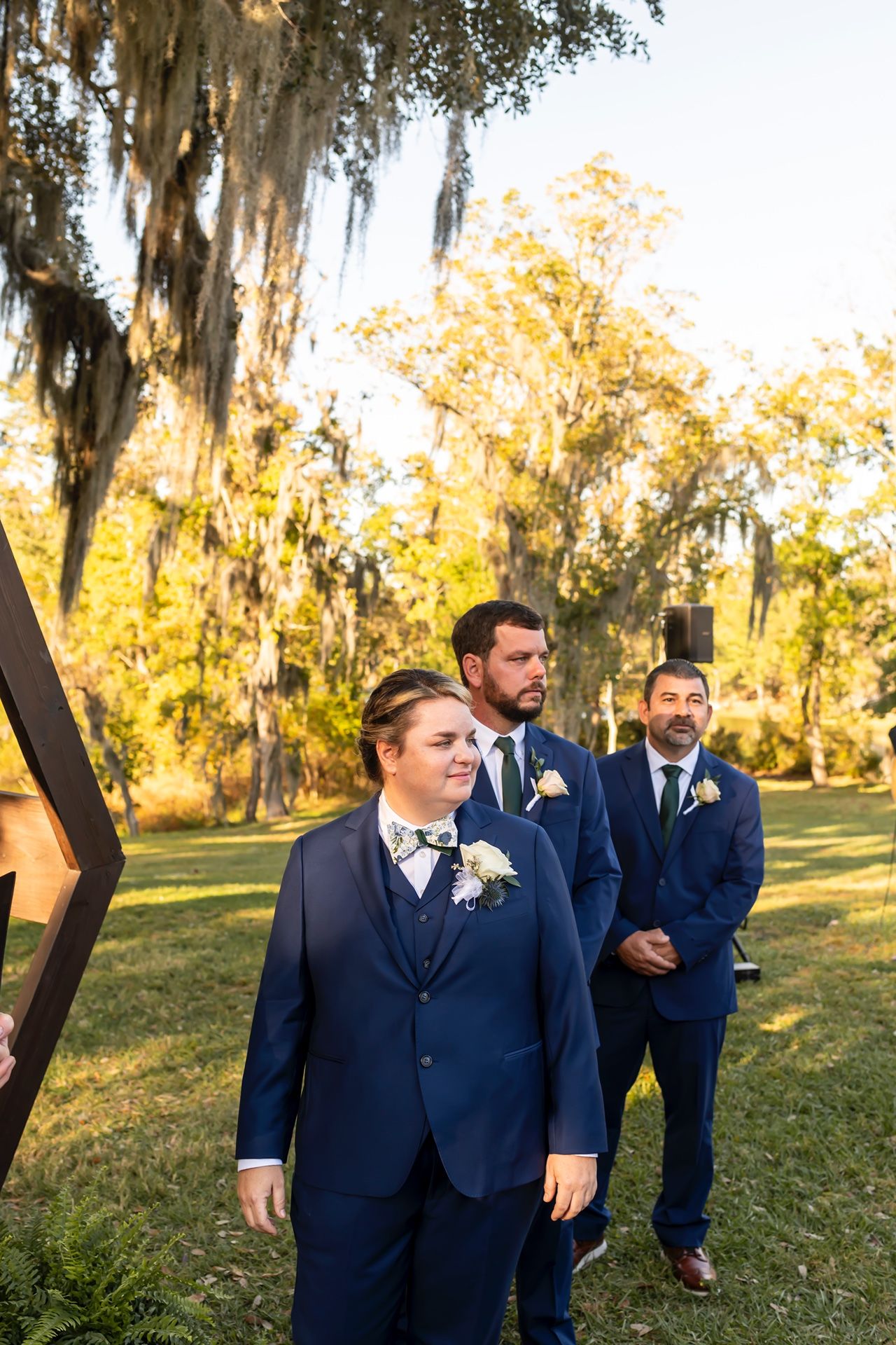 Three people in navy blue suits stand outdoors on a sunny day with Spanish moss draped trees in the background.