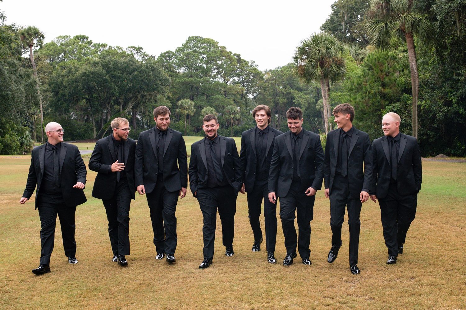 A line of groomsmen in black suits walking together on grass during an outdoor wedding.