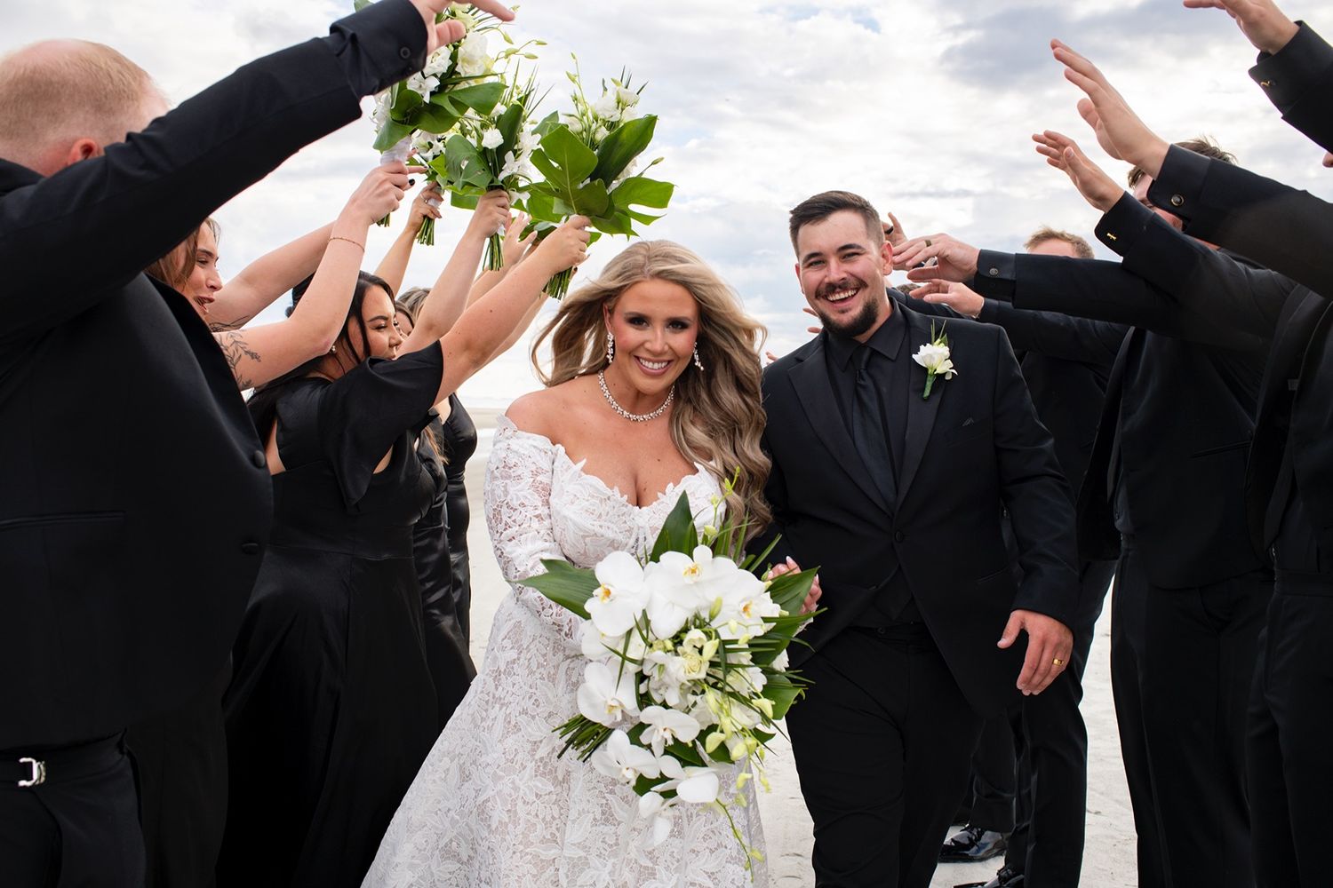 Wedding party forms a tunnel of raised arms as newlyweds walk through holding white floral bouquet at outdoor celebration.
