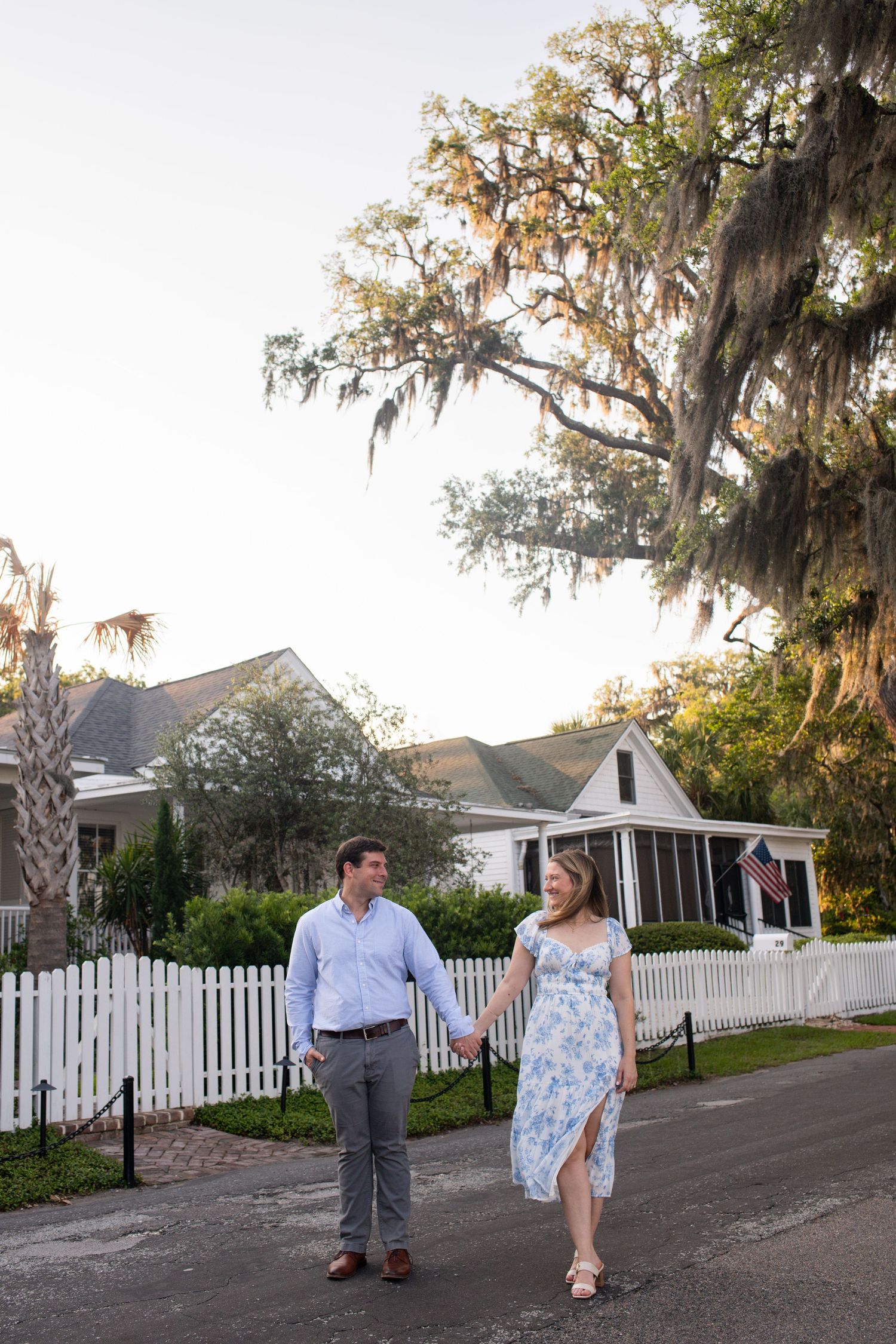 Couple walks together on a pathway with Spanish moss draped trees and white picket fences in a romantic garden setting.