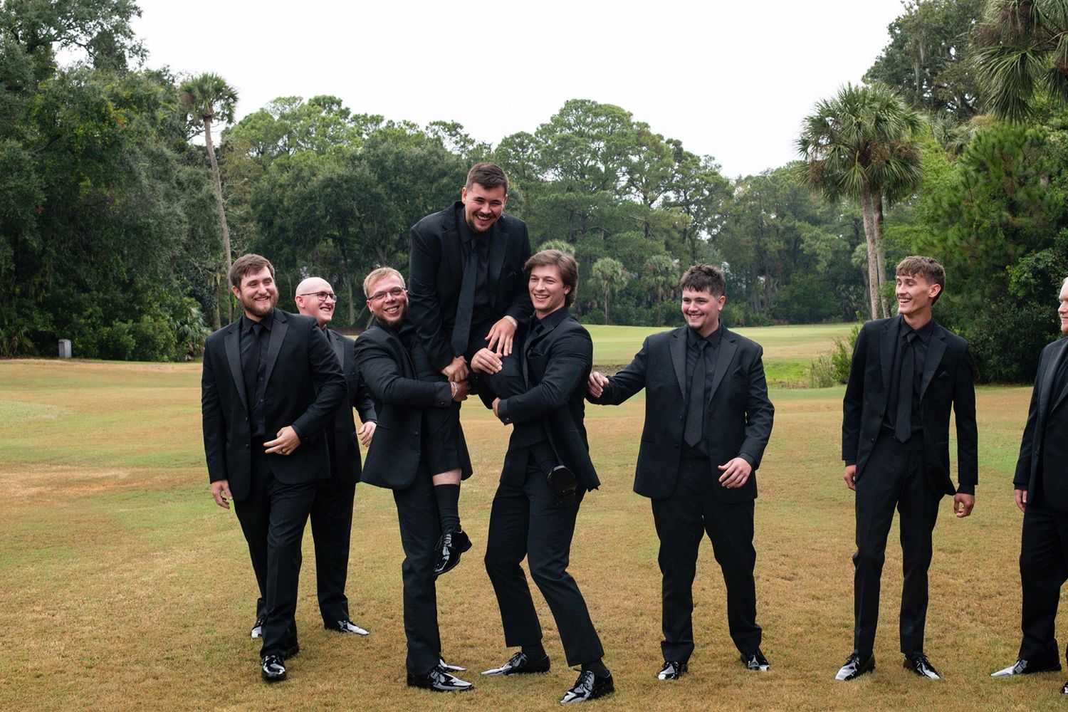 Group of people dressed in all black outfits standing together and posing playfully on a grassy lawn with trees.