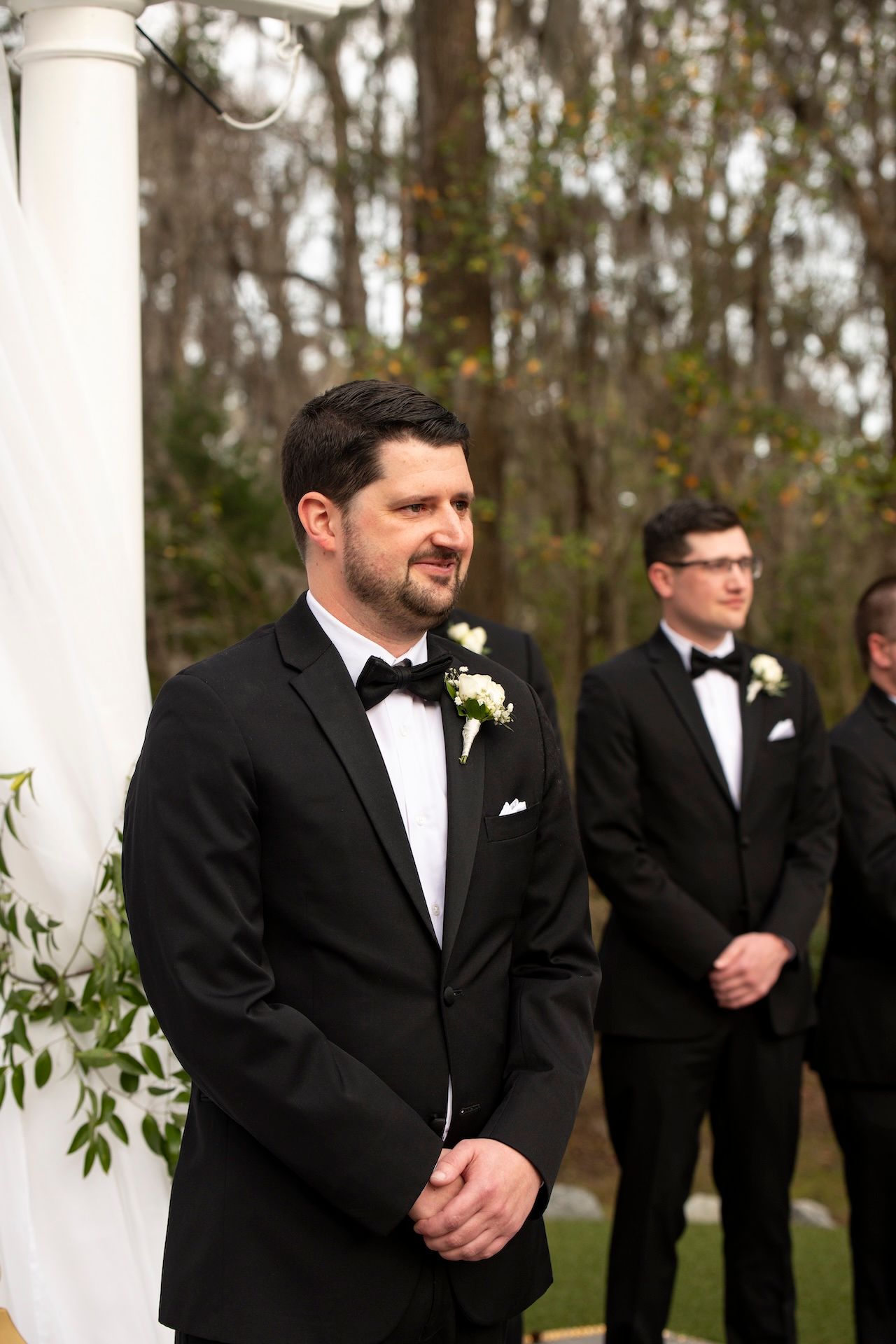 Groom in black tuxedo with white boutonniere smiles at altar during outdoor wedding ceremony with groomsmen behind him.