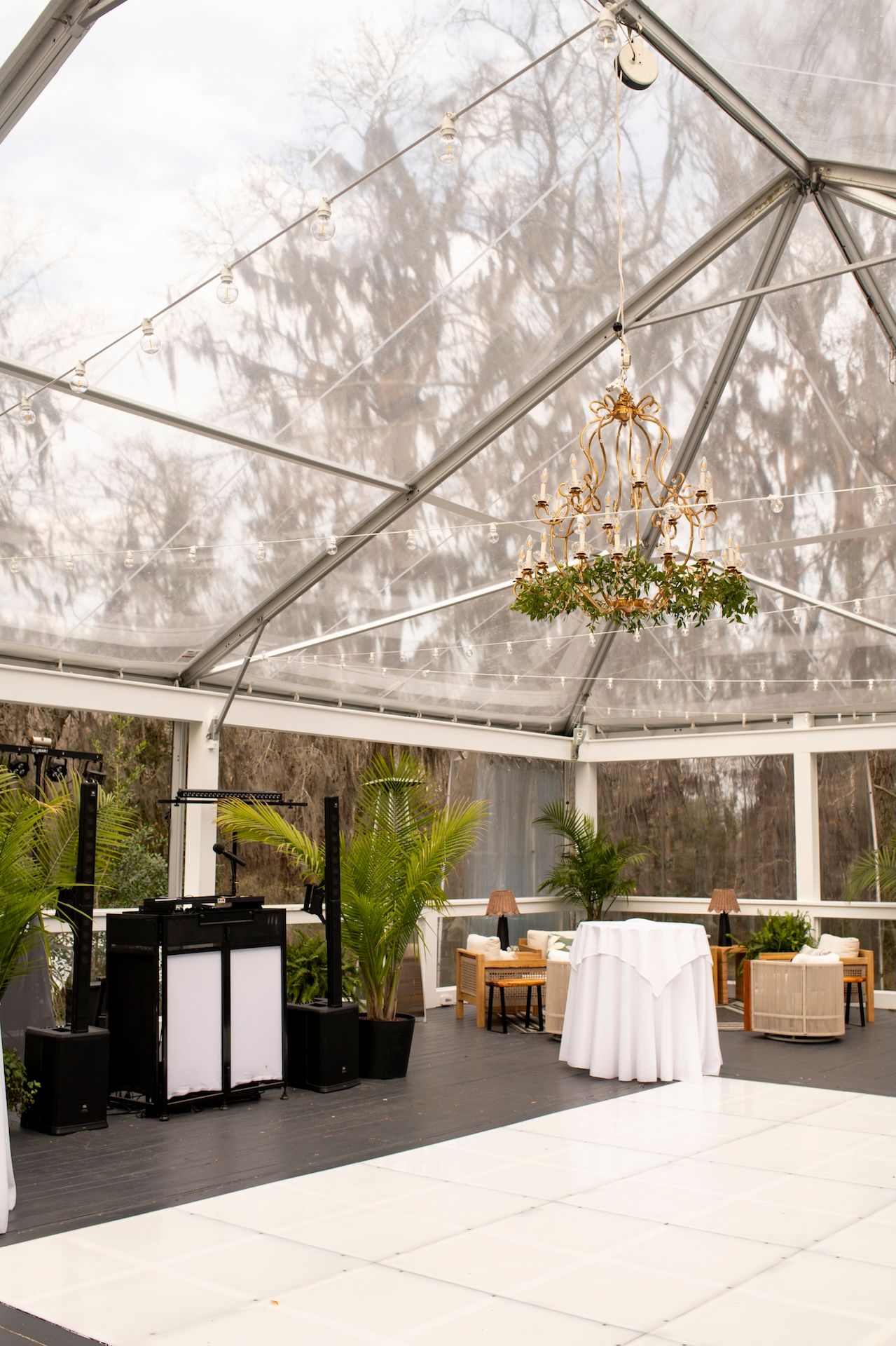 Elegant clear tent venue with chandelier, white draped tables, fern decor, and DJ setup on white dance floor.