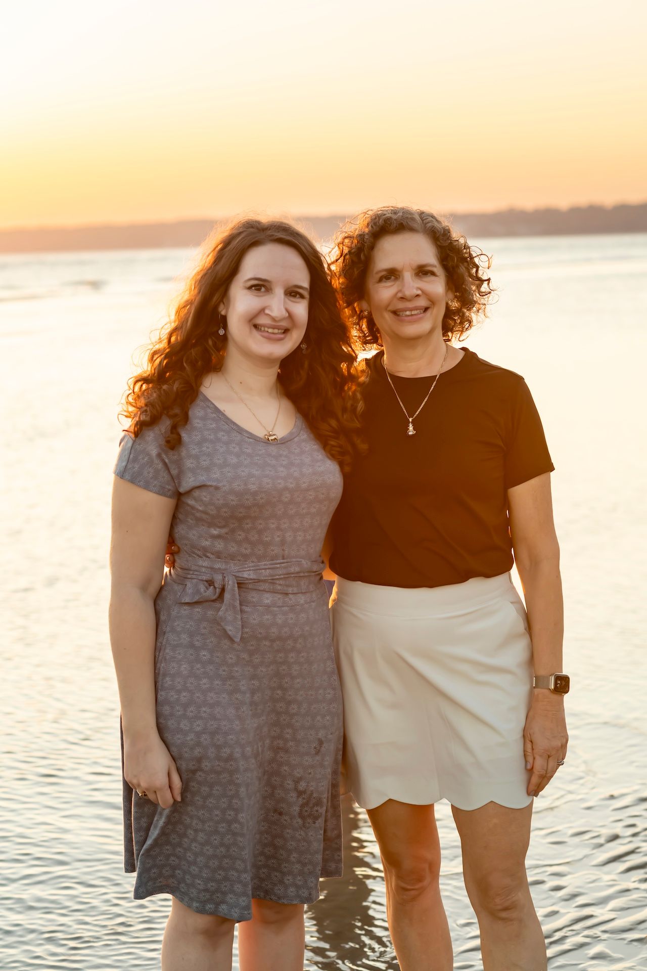 Two people in casual attire standing together at sunset by the water with a warm glowing sky in the background.