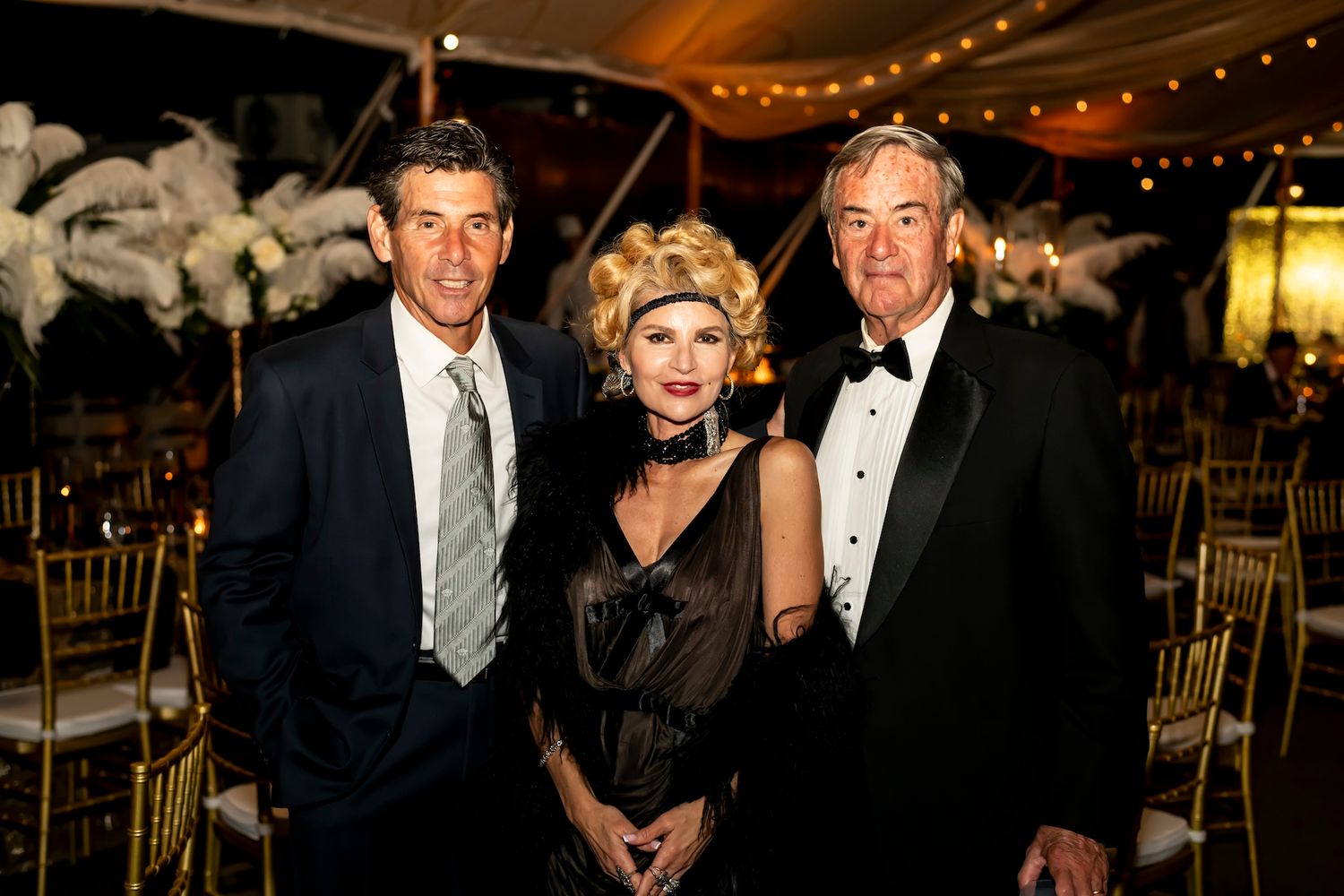 Three people pose together at a formal evening event with string lights and decorations visible in the background.