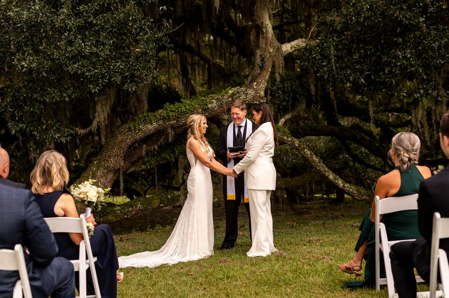 An outdoor wedding ceremony takes place under mossy oak trees as guests watch from white chairs in a garden setting.