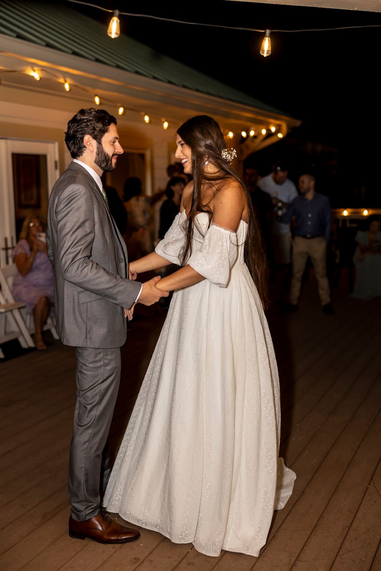 A couple shares a romantic moment on a wooden deck under string lights at an evening wedding celebration.