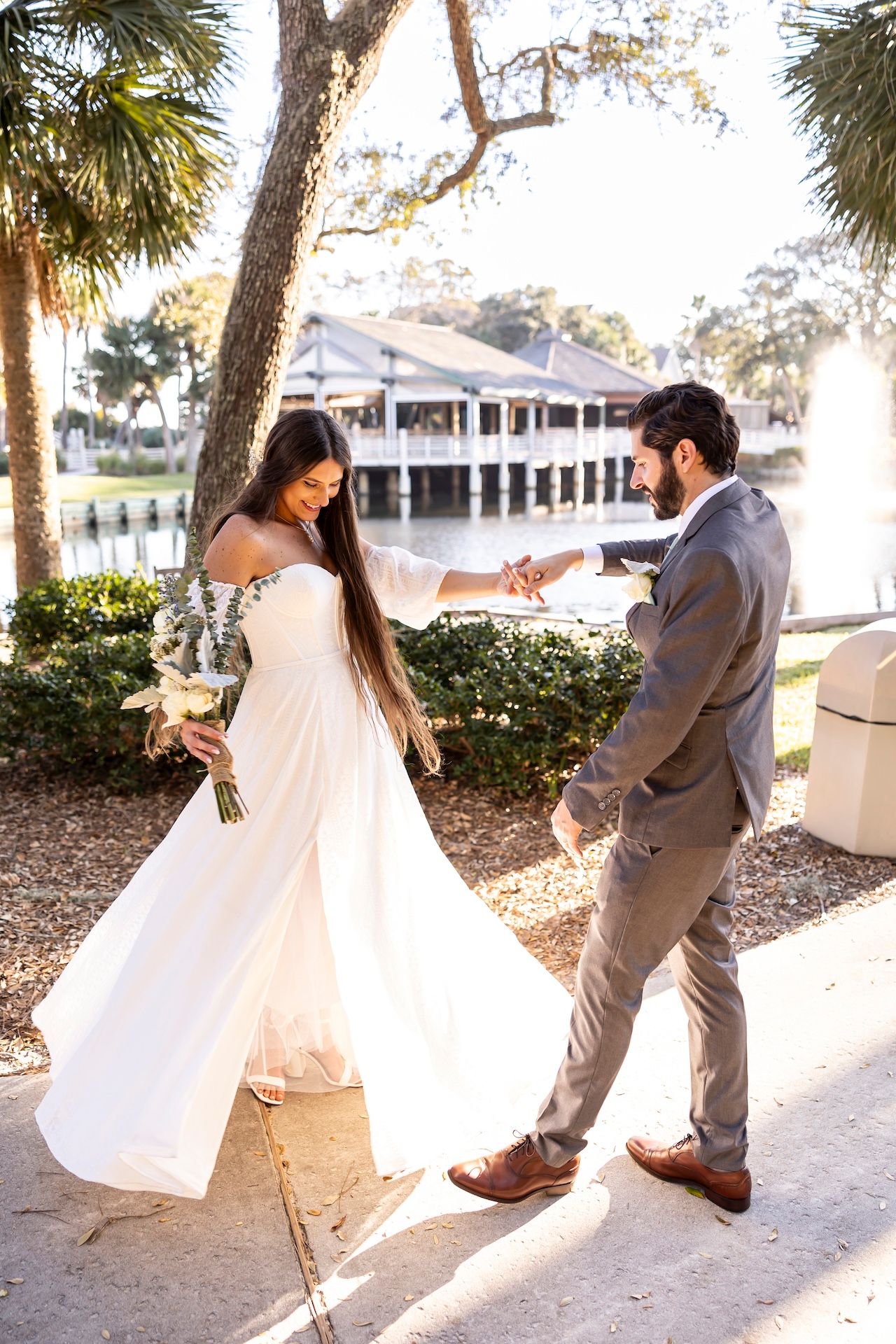 Beautiful wedding couple shares romantic moment at sunset by lakeside venue with palm trees and white building.