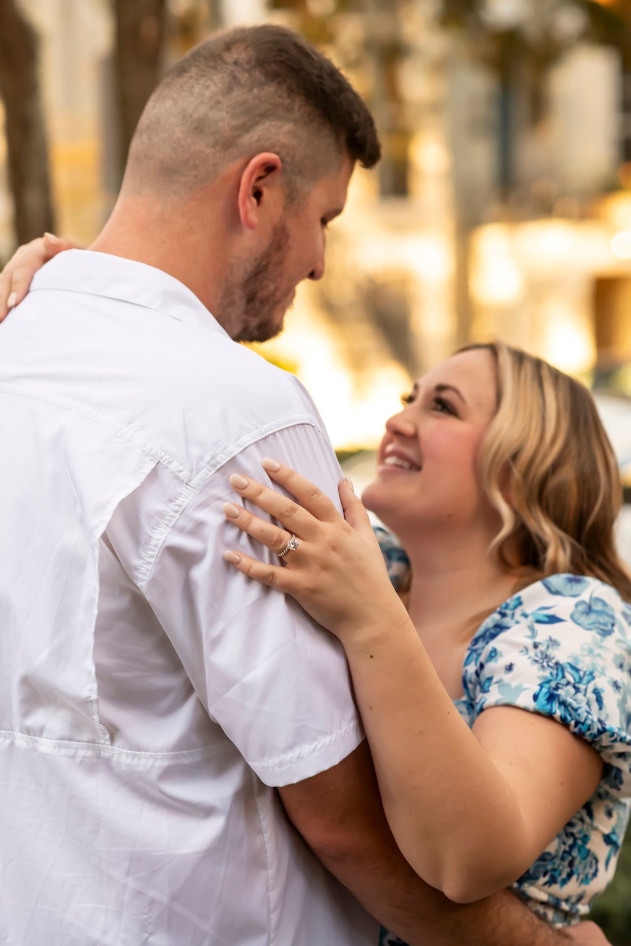 A romantic couple embraces outdoors, with the woman wearing a floral dress and smiling while touching her partner's arm.