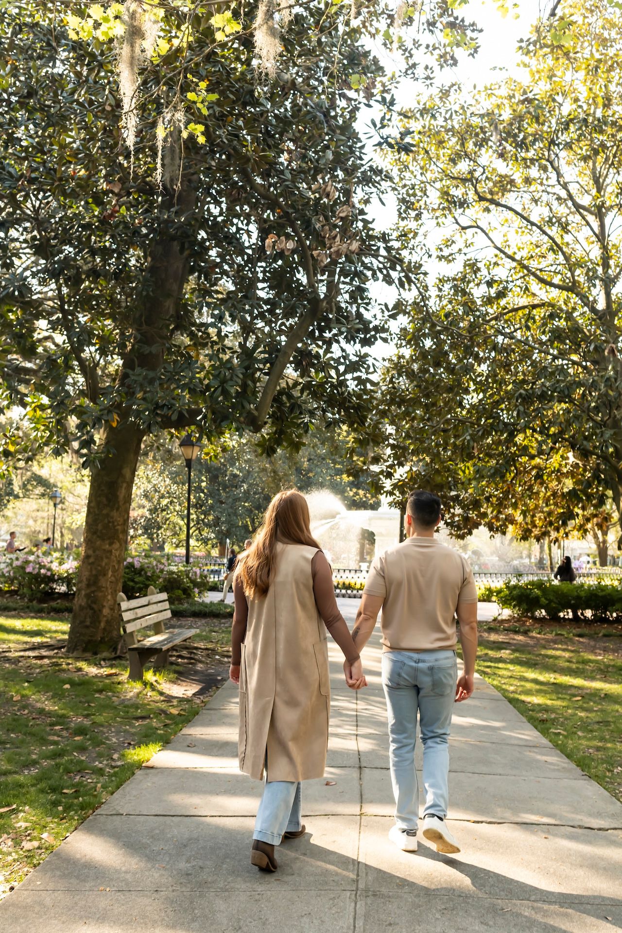 A couple walks hand in hand down a sunlit path surrounded by autumn trees in a park.