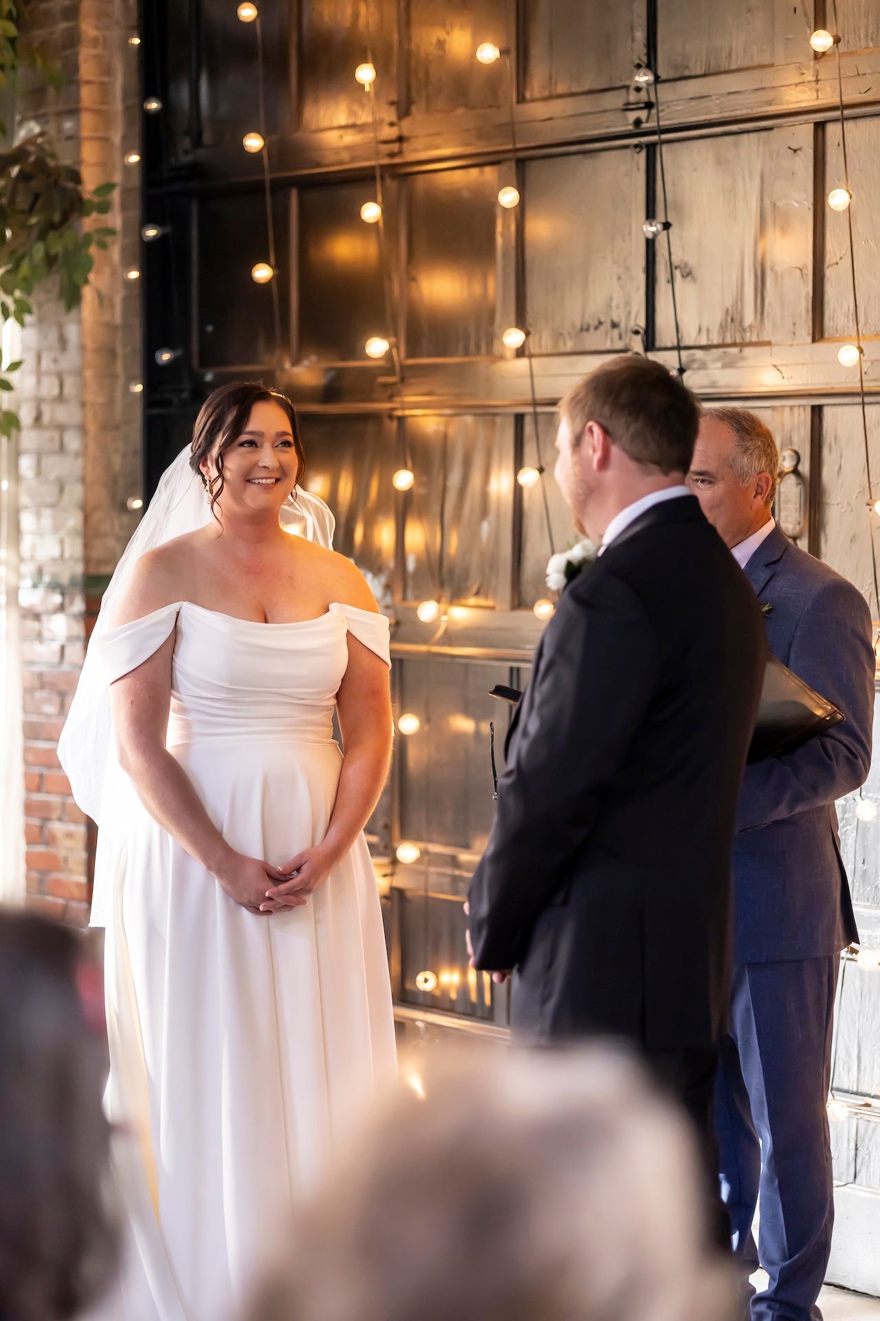 A couple exchanges vows during their wedding ceremony in an industrial-chic venue with warm string lights and exposed brick.