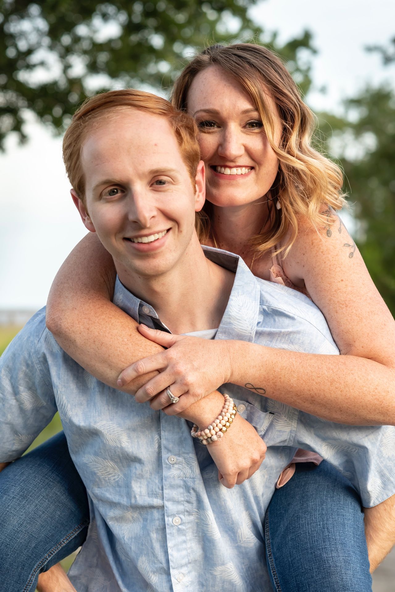 A couple shares a joyful moment outdoors with a piggyback pose against a blurred natural background.