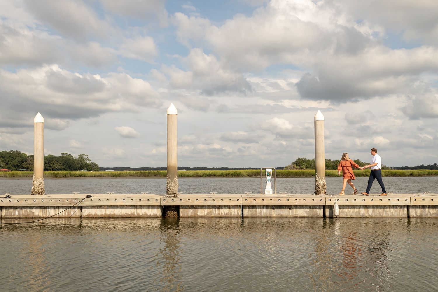 People walking along a coastal boardwalk with white poles and railings under cloudy skies near the water's edge.