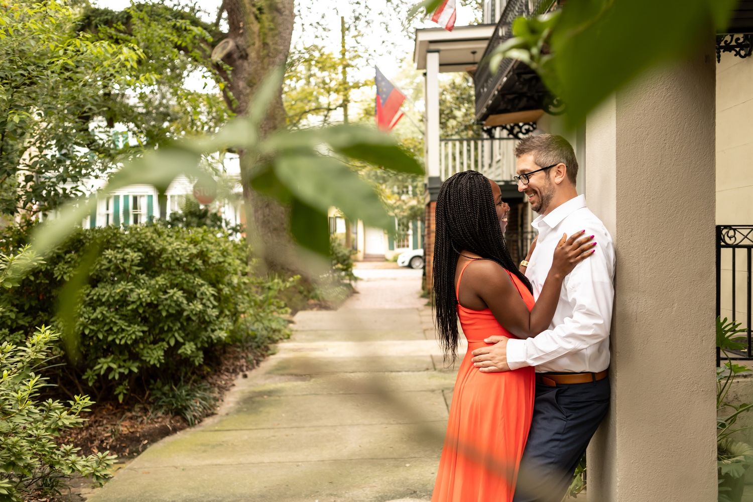 A couple embraces on a tree-lined sidewalk near a white building on a sunny day.