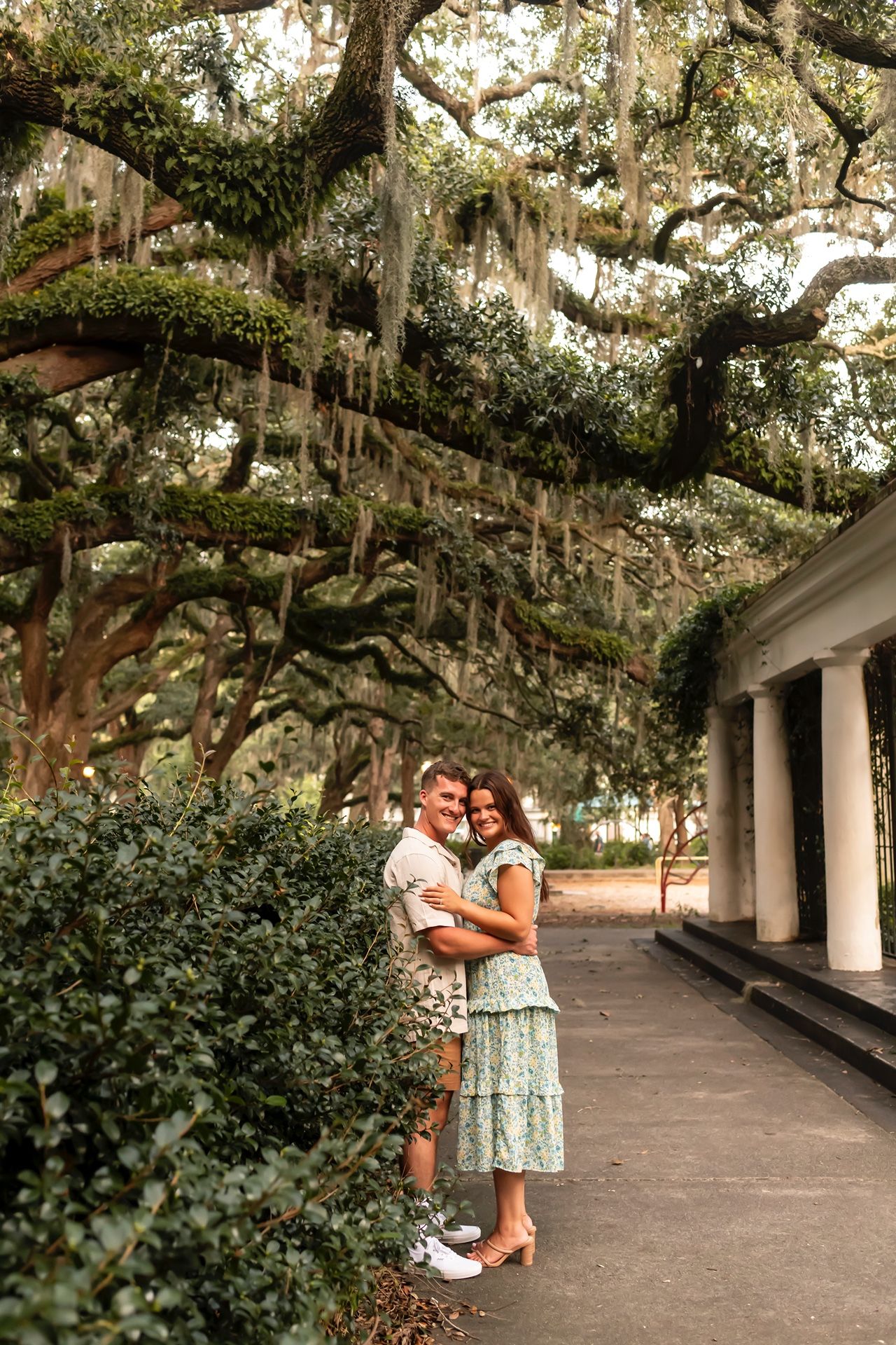 A romantic couple embraces under Spanish moss-draped oak trees beside a columned building walkway.