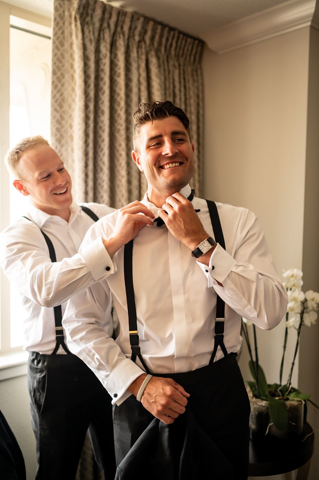A groom gets help adjusting his suspenders while getting ready in a well-lit room with floral decor.