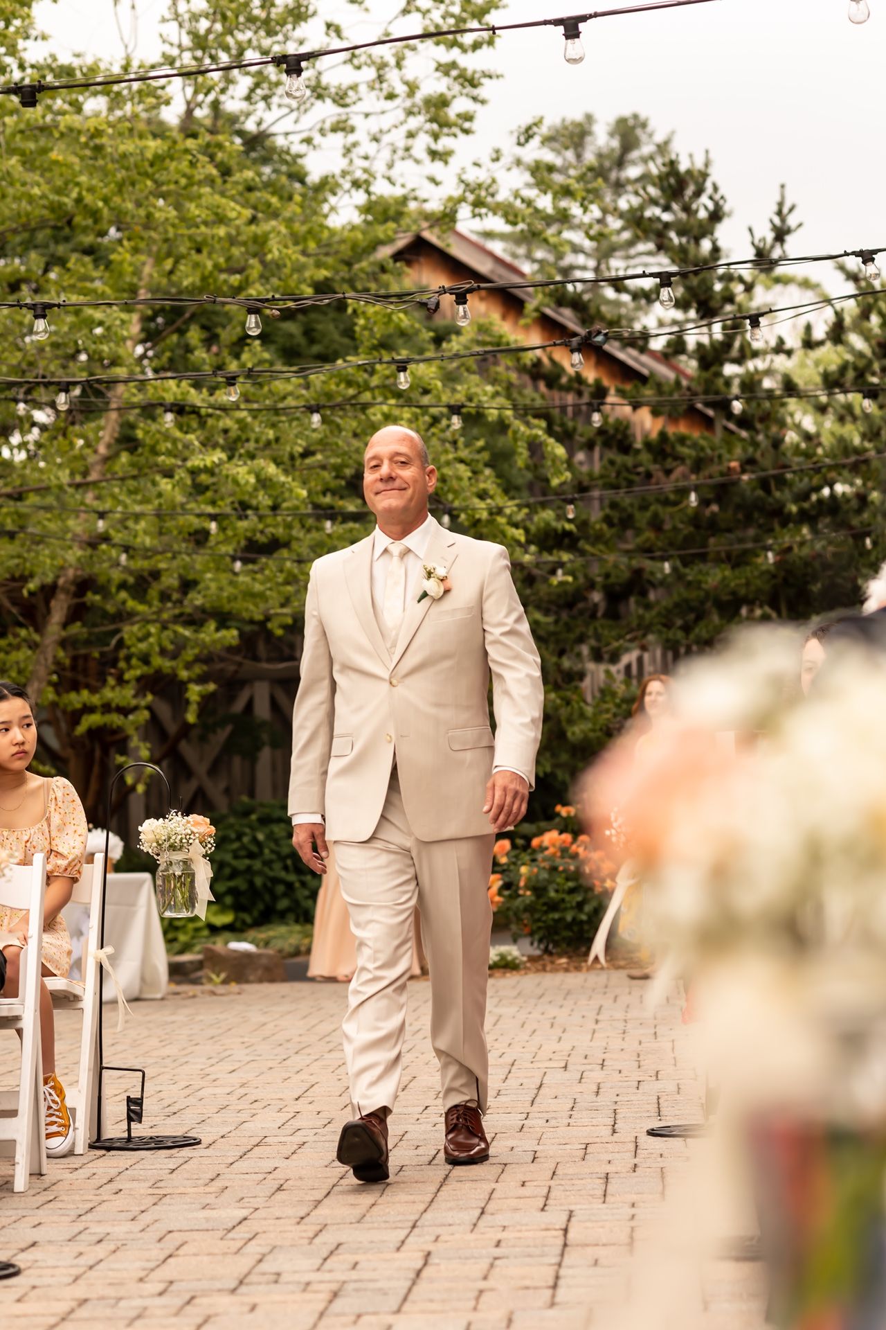 A wedding guest in a tan suit walks down a stone paved path at an outdoor ceremony venue.
