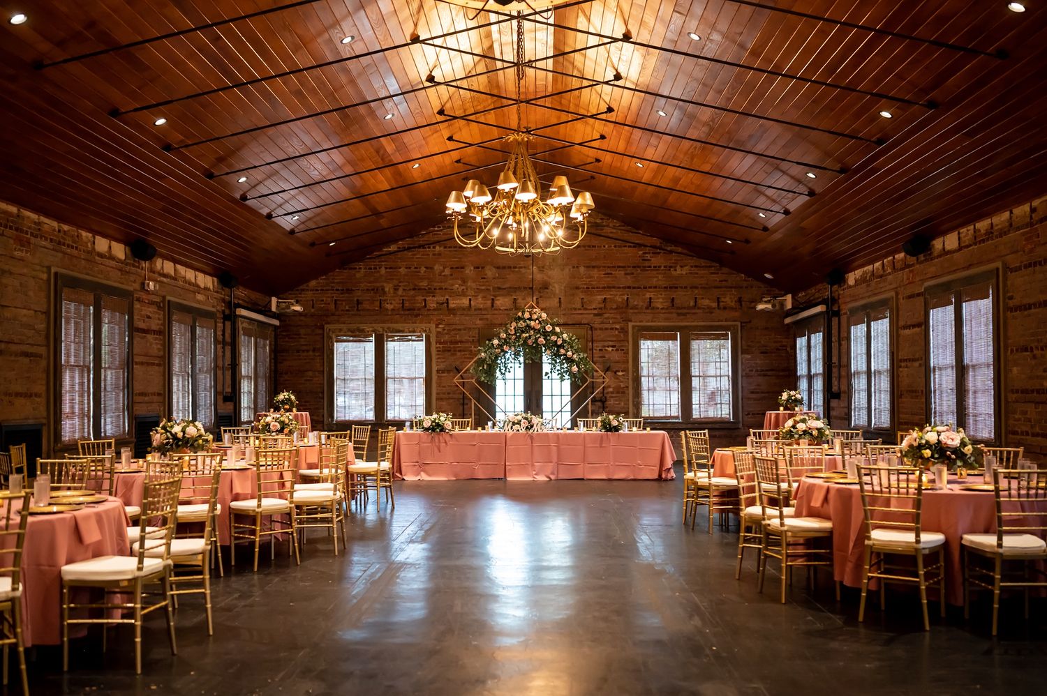 Elegant rustic wedding reception venue with wooden beams, chandelier, and tables set for dining in warm ambient lighting.