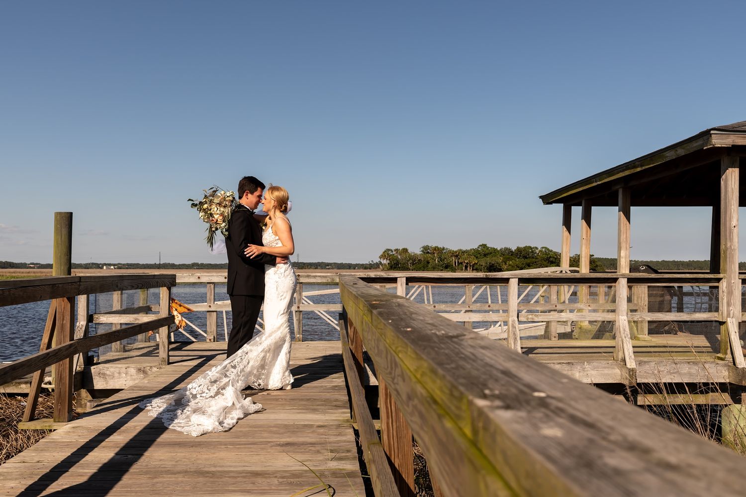 Newlyweds share an intimate moment on a wooden dock overlooking a scenic coastal marsh at sunset.
