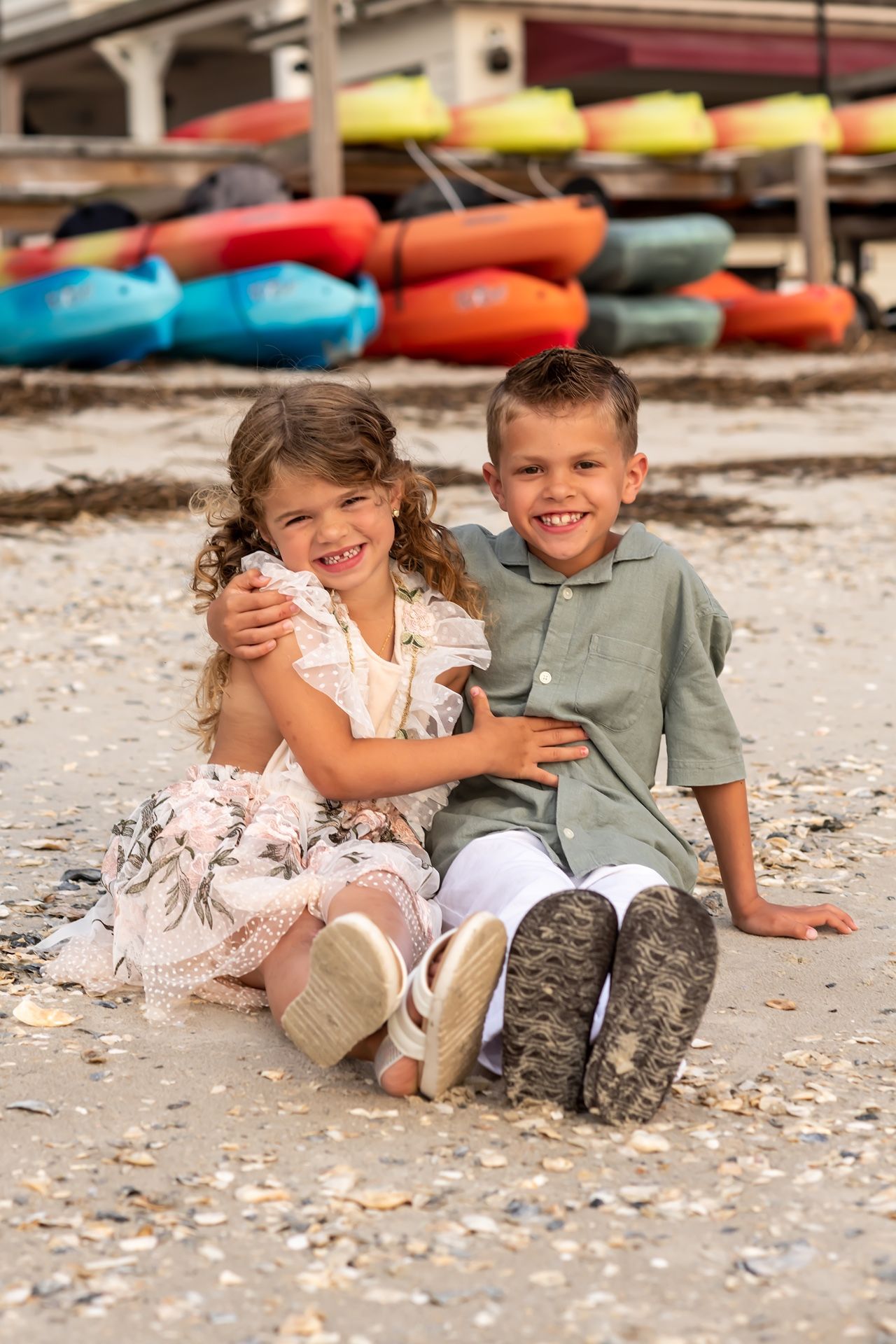 Two children in casual clothes sitting together and laughing on a dock with colorful kayaks behind them.
