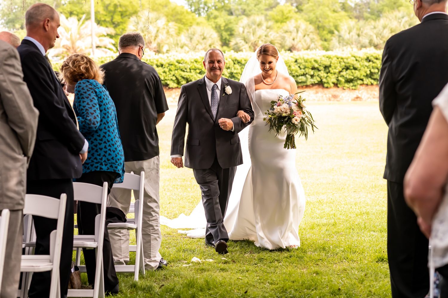 A couple walks down a grassy outdoor aisle surrounded by wedding guests seated in white chairs on a sunny day.