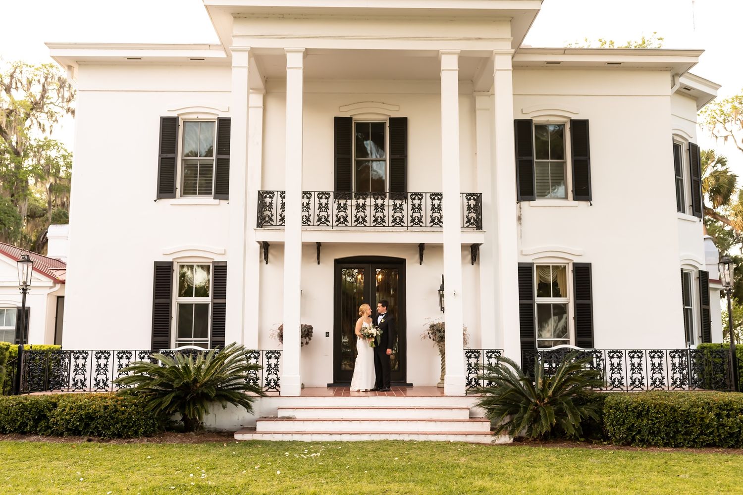Two figures stand on the grand front porch of a classic white Southern antebellum mansion with towering columns and black shutters.