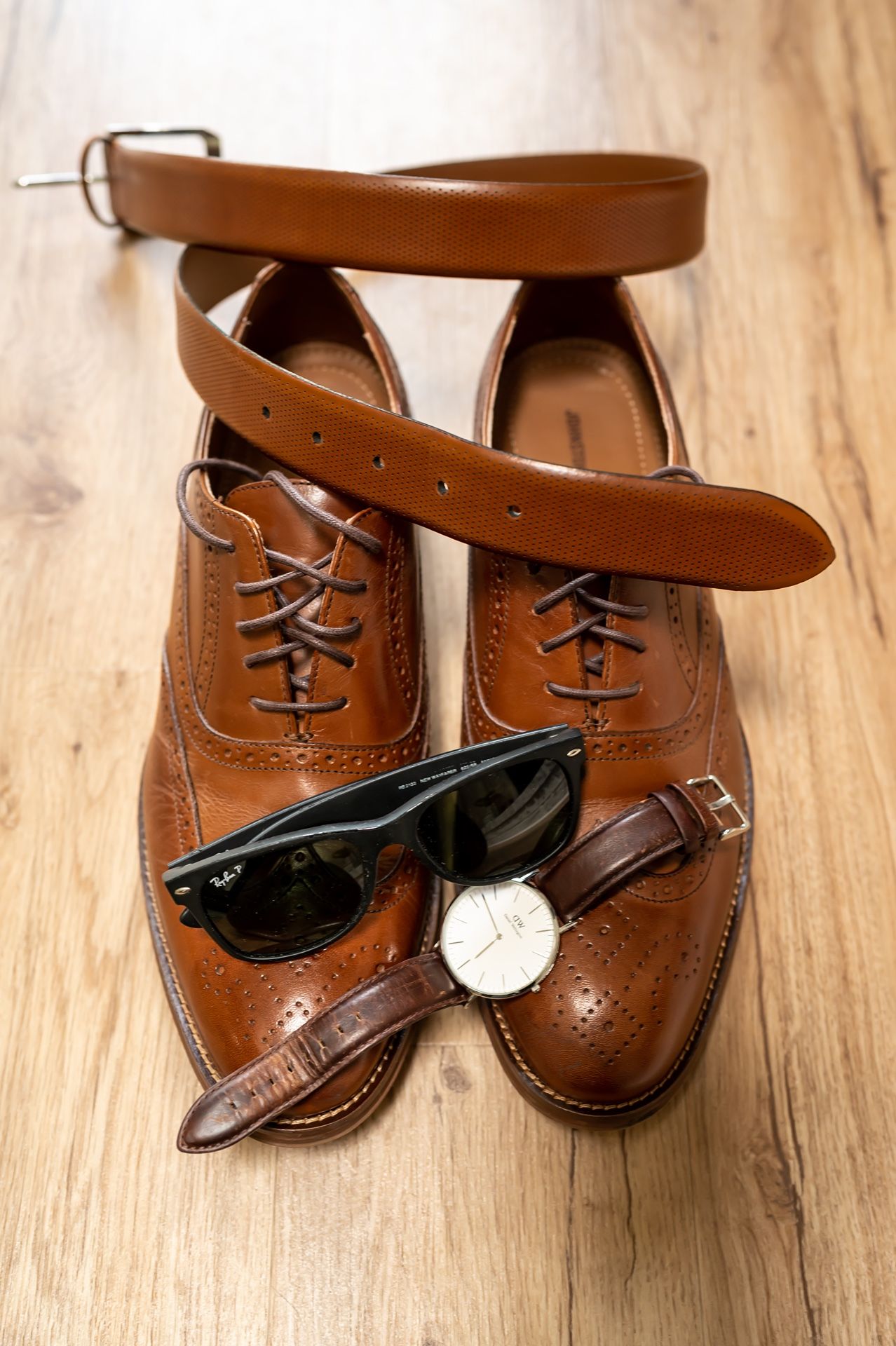 Brown leather oxford dress shoes arranged with leather belt, sunglasses and wrist watch on wooden floor.
