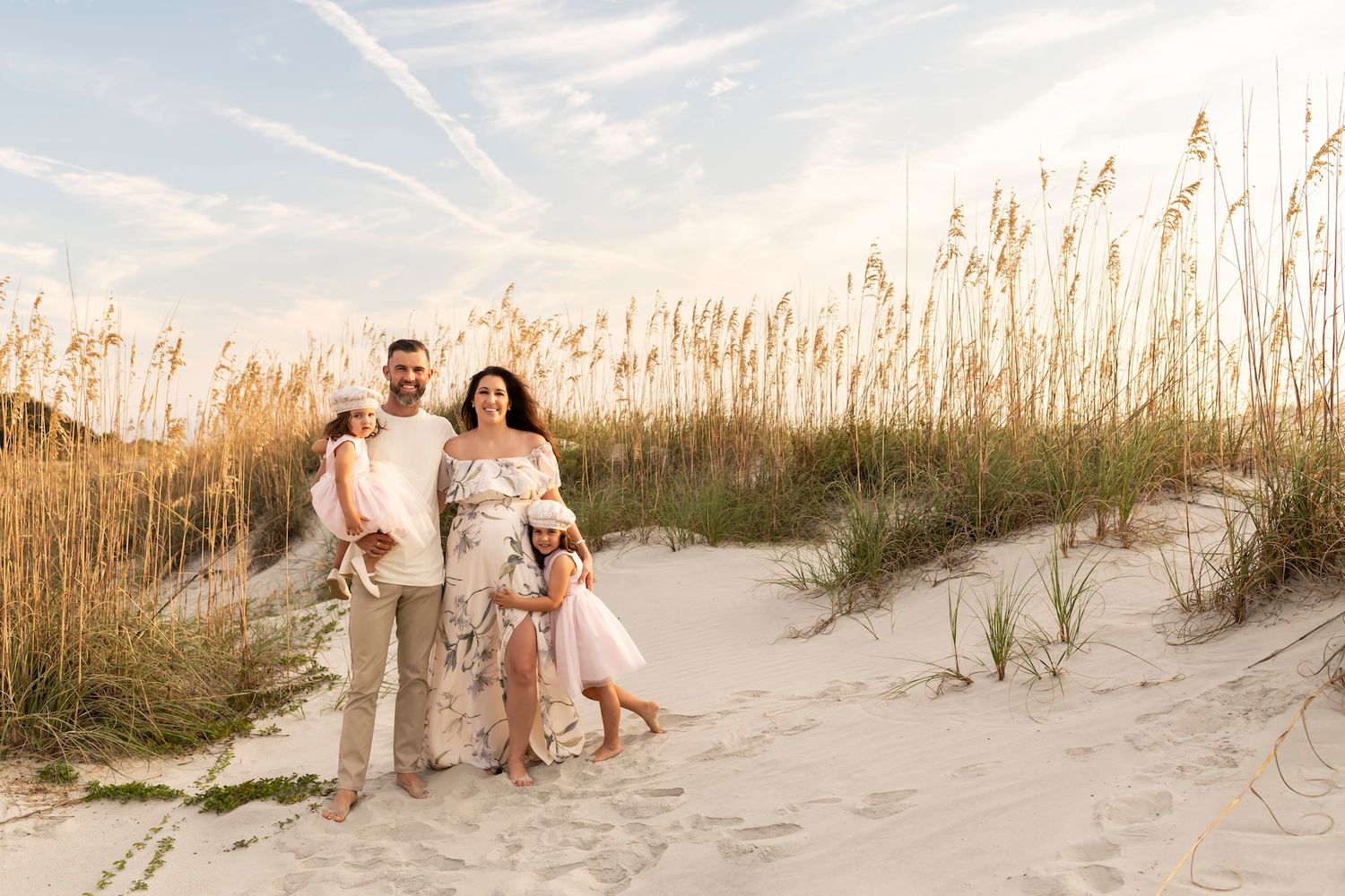 A couple and their dog stand together on a sandy beach path surrounded by tall grass and dunes at sunset.