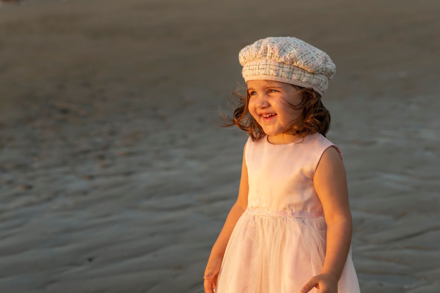 A child in a white summer dress and matching cap stands on a beach at golden hour.