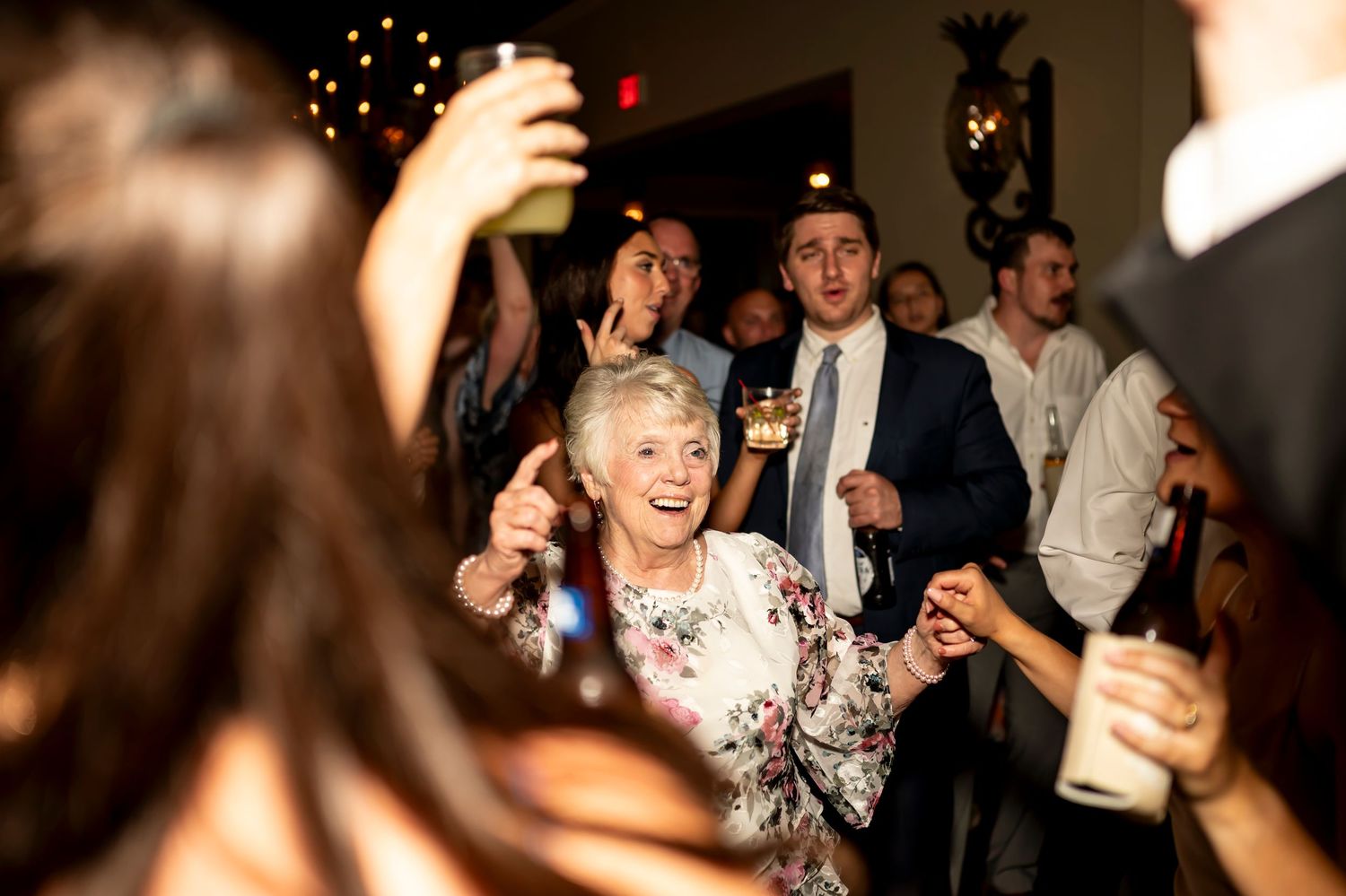 Party guests laughing and dancing while raising drinks in celebration at a festive indoor event with twinkle lights.