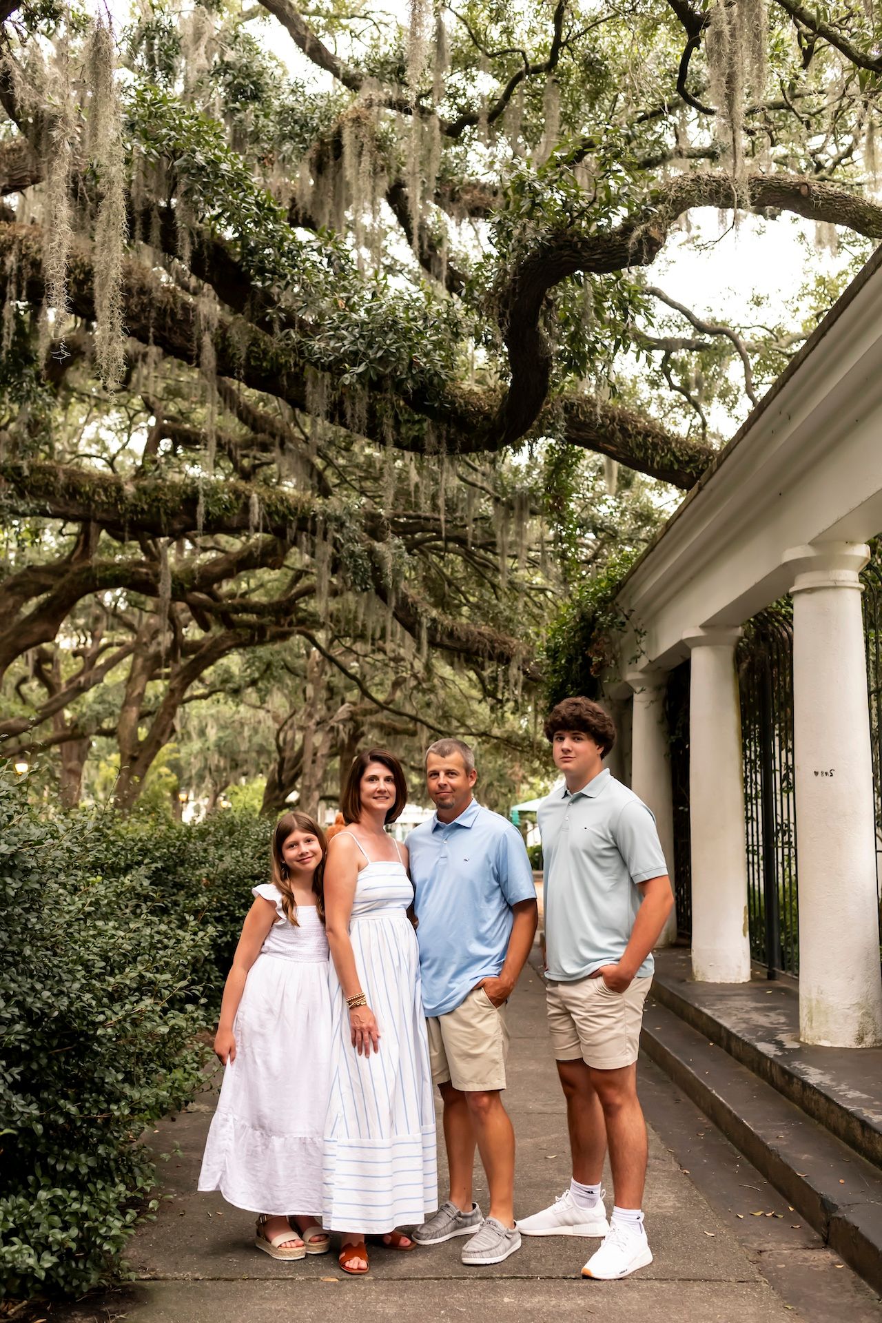 Family group poses under Spanish moss draped trees near white columns of a Southern building.