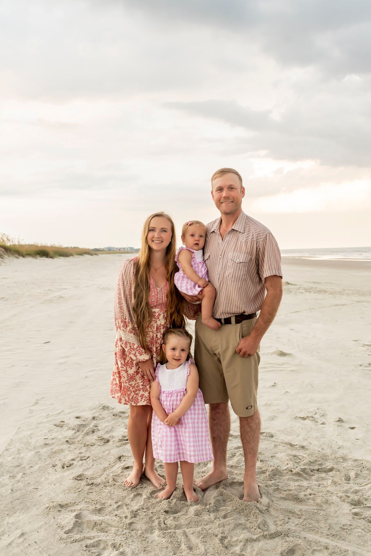 A family stands together on a beach at sunset with sand dunes and ocean waves visible in the background.