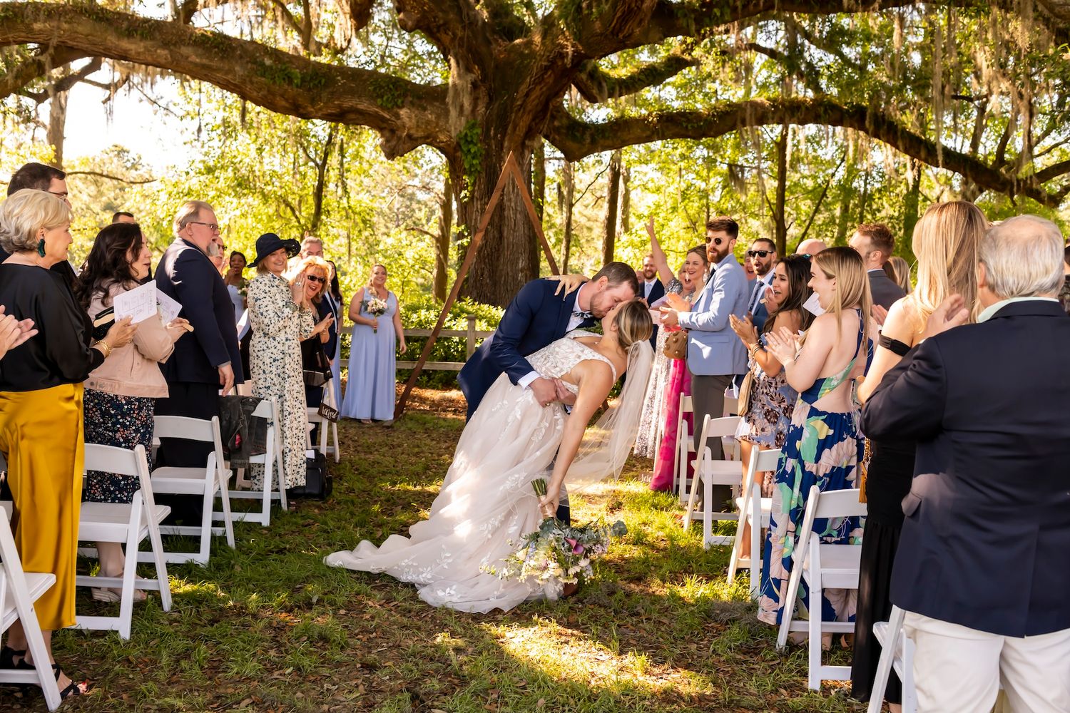 Outdoor wedding ceremony under a rustic wooden pergola with rows of white chairs set up on a grassy lawn in natural sunlight.