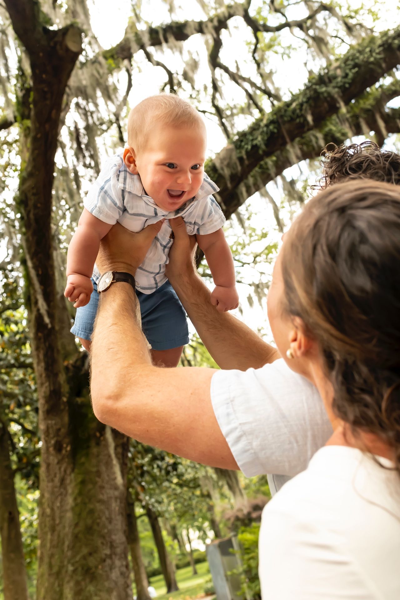 A baby laughs with joy while being lifted up by someone under Spanish moss draped trees.