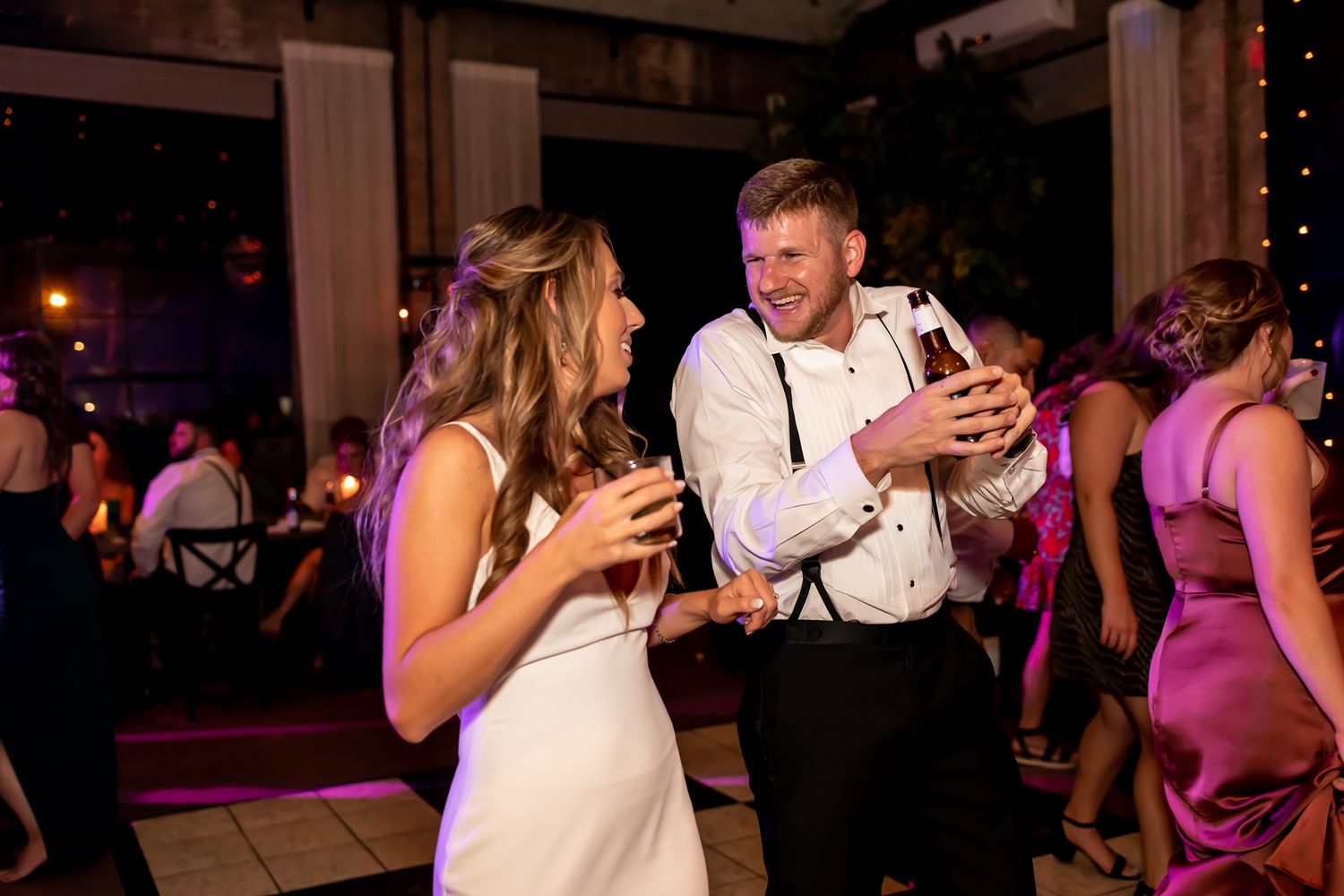 Guests celebrating on a crowded dance floor at an evening wedding reception with purple ambient lighting.