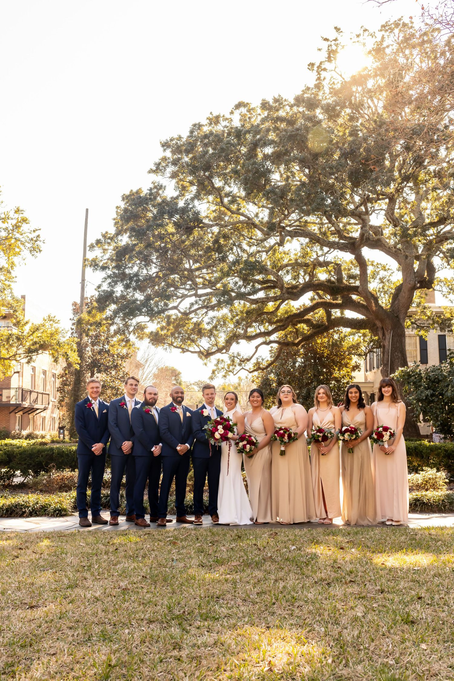 Wedding party poses outdoors under large oak tree at sunset with bridesmaids in champagne dresses and groomsmen in navy suits.