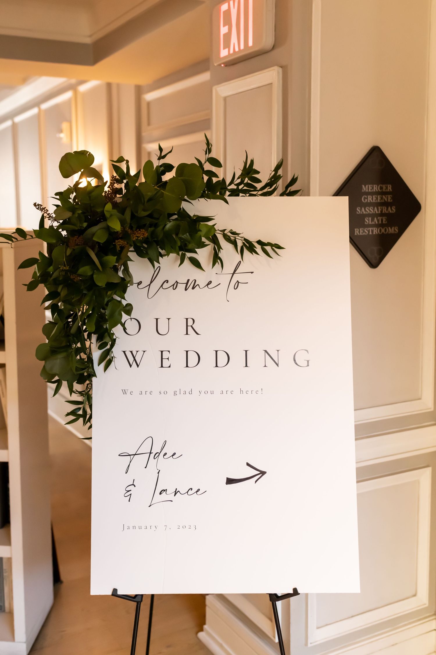 Minimalist wedding signage with greenery decorations hangs on a white wall near an exit sign.